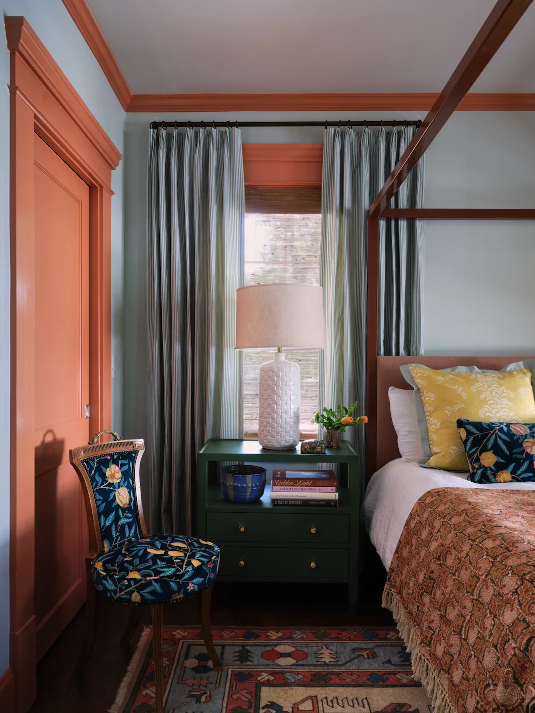 A bedroom features light blue walls with coral-painted trim, a dark green nightstand, striped curtains, and a patterned botanical side chair.