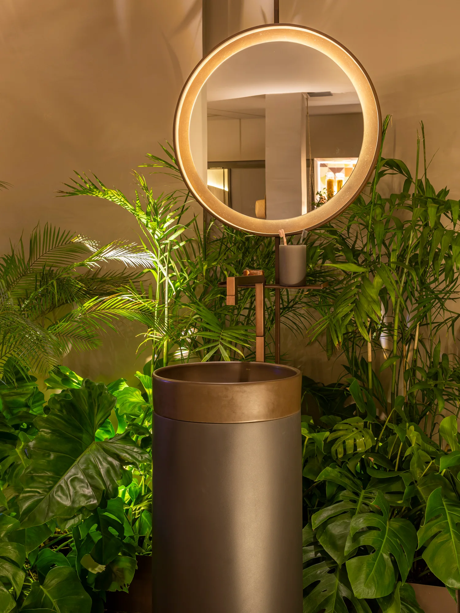 A modern gray cylindrical pedestal sink and a glowing backlit circular mirror are surrounded by dense green potted foliage.
