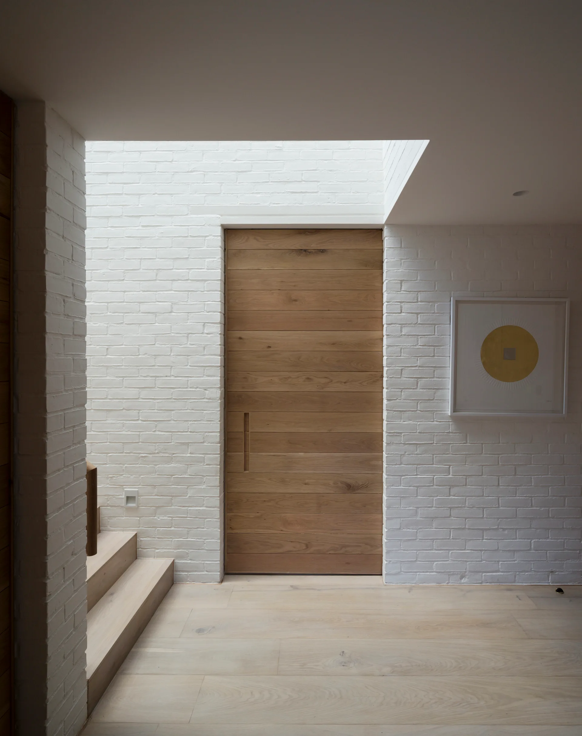 A minimalist entryway features white painted brick walls, a natural wood door, a skylight overhead, and light wood floors.