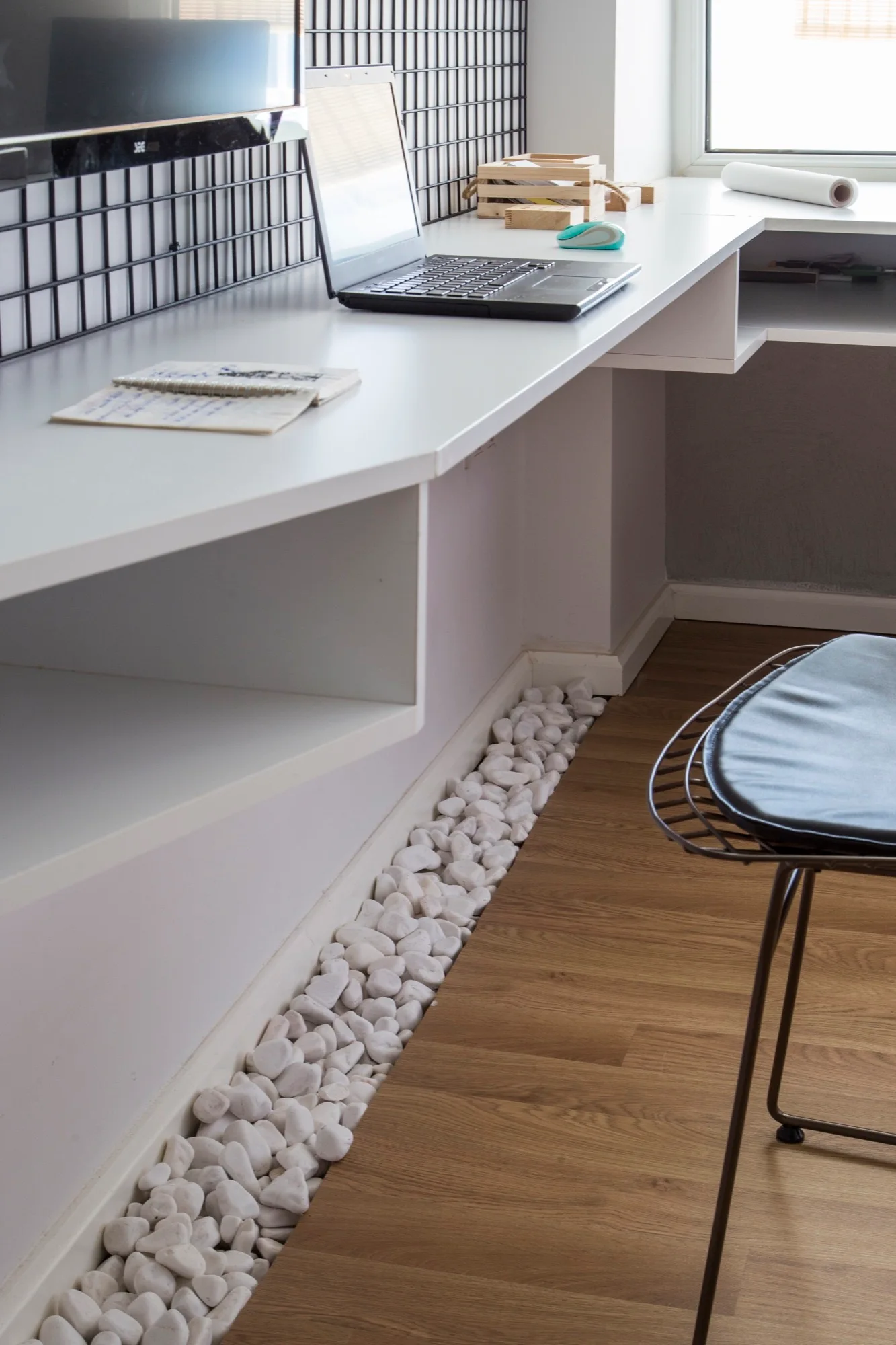 A white floating desk in a home office features a laptop, a black wire wall grid, and white decorative pebbles lining the wooden floor below.