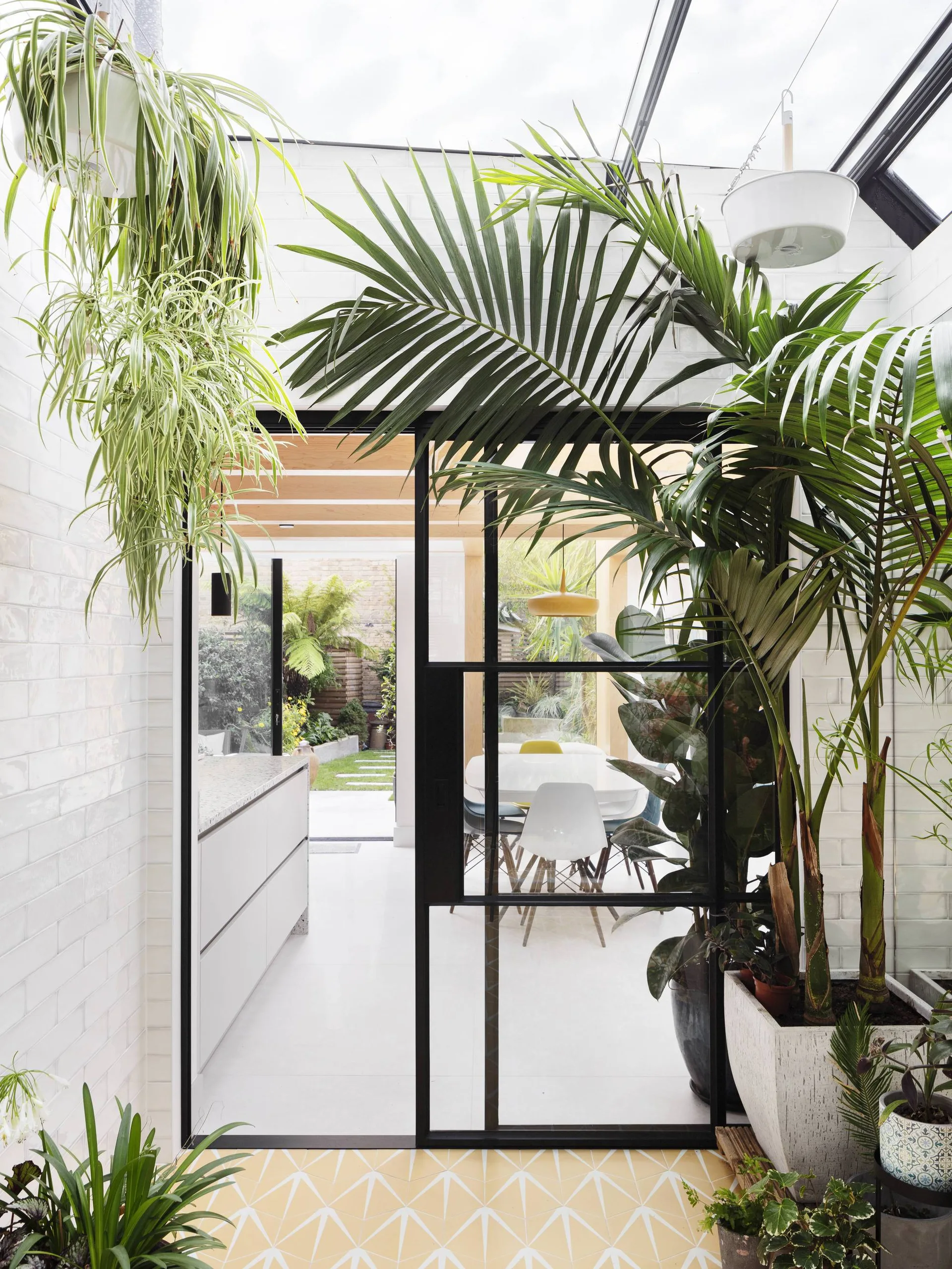 A black-framed glass sliding door separates a sunroom with yellow patterned tile flooring and potted palms from a white kitchen and dining area.