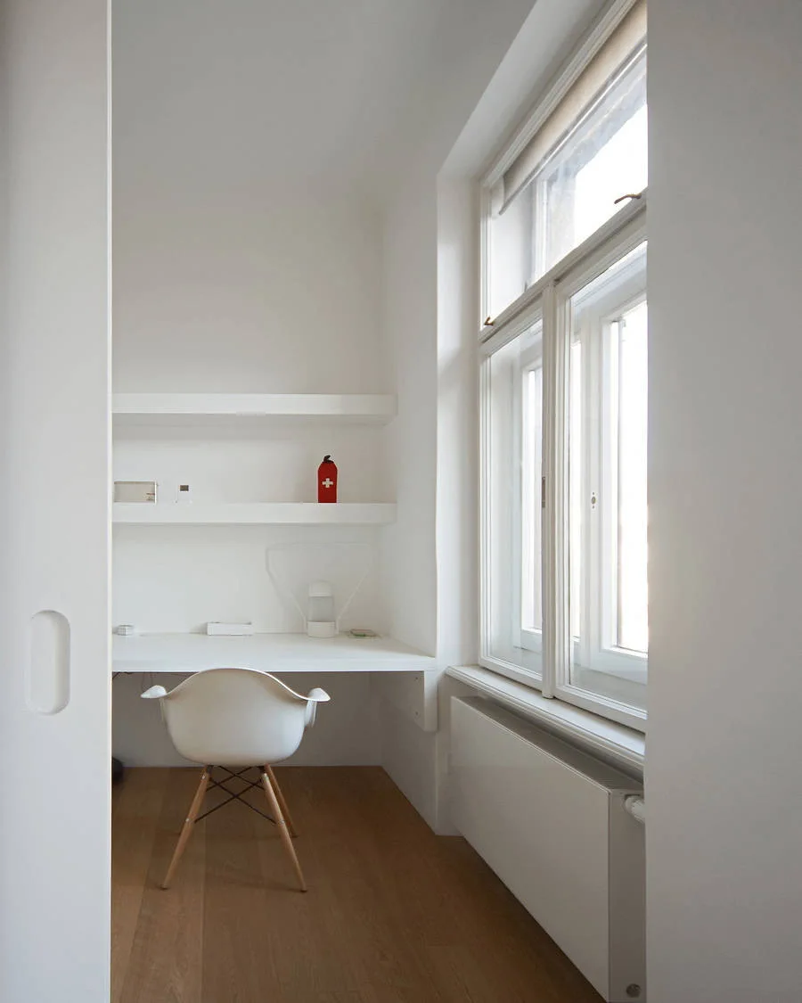 A minimalist white home office nook features a built-in desk, floating shelves, a white chair with wooden legs, and a large window.