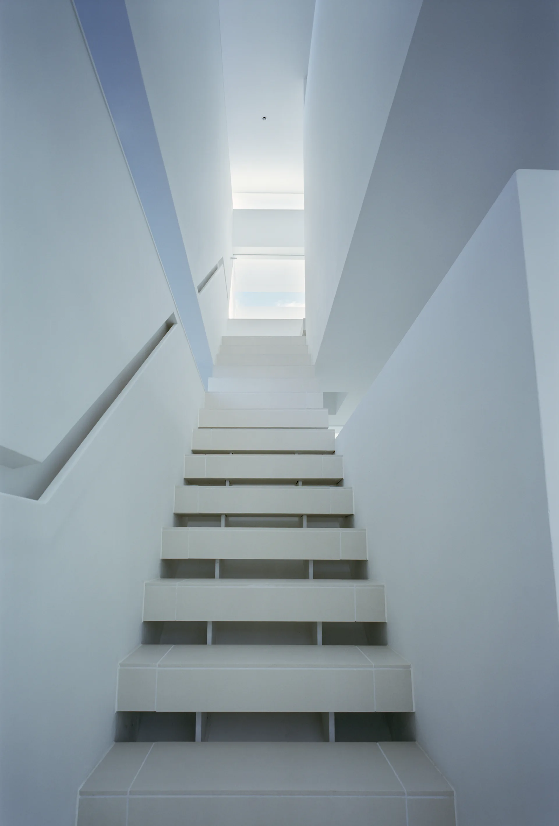 A minimalist white staircase constructed of simple tiled steps ascends between smooth white plaster walls toward a bright, sunlit overhead opening.