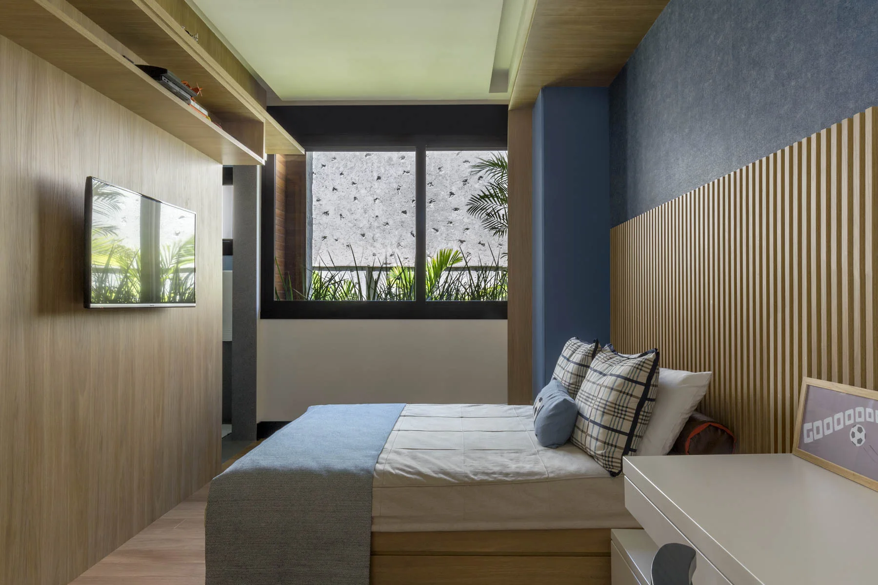 A contemporary bedroom features a bed with plaid pillows set against a wood-slatted headboard wall, opposite a wood-paneled wall with a mounted TV.