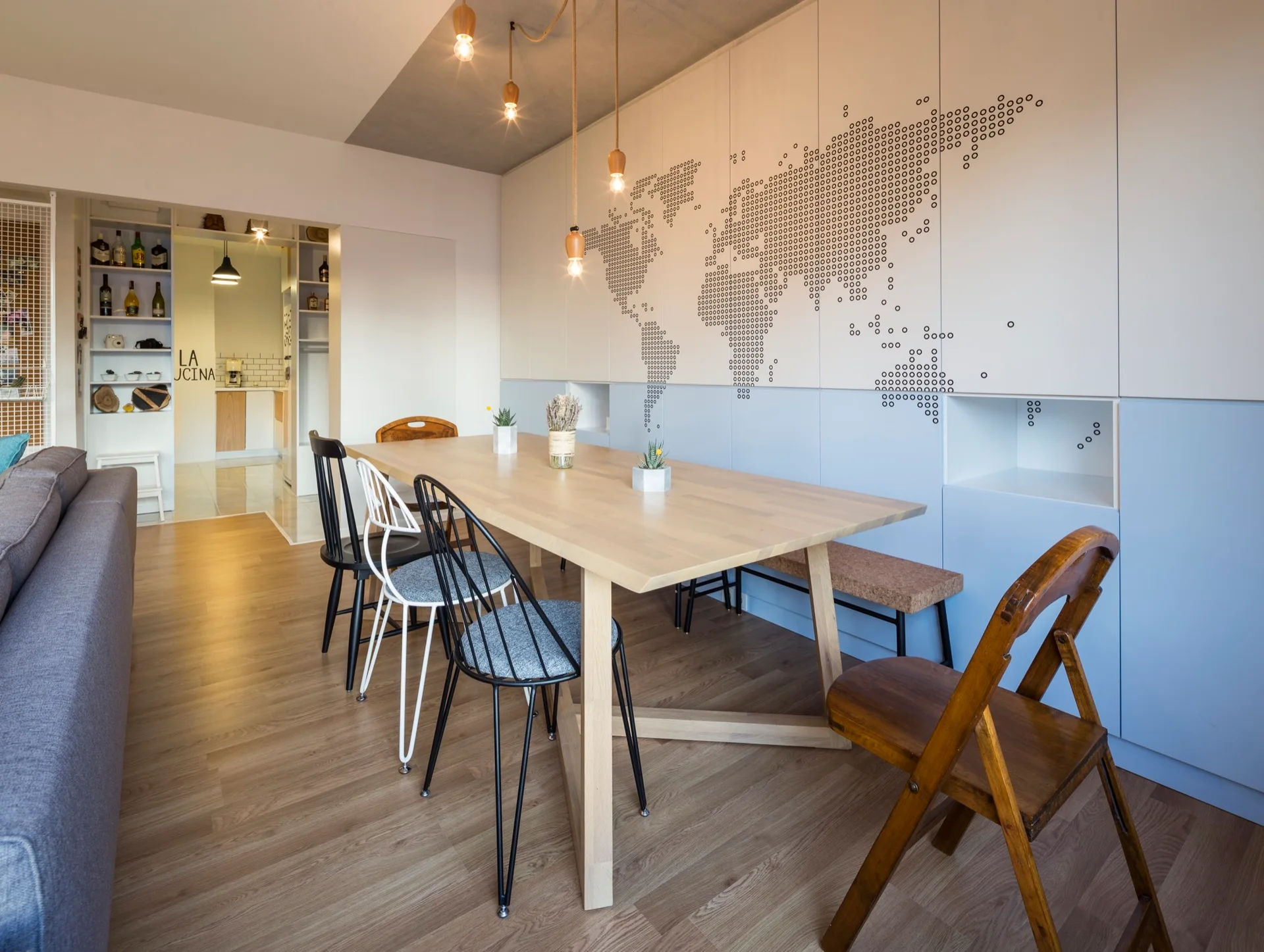 A light wood dining table with mismatched chairs stands before a large white and blue cabinet featuring a dotted world map design.