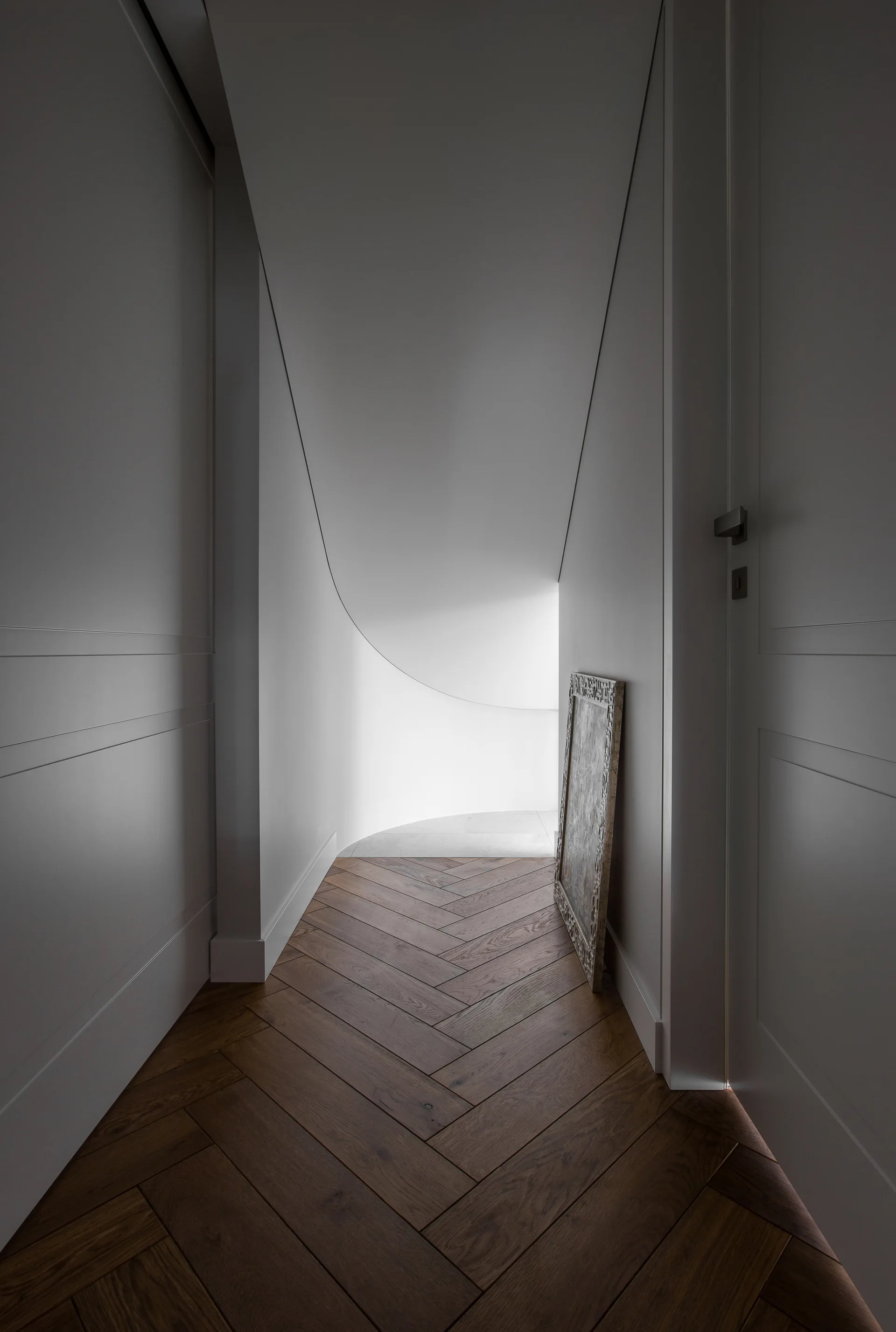 A narrow white hallway with dark herringbone wood flooring, paneled doors, and a curved architectural transition leading to natural light.