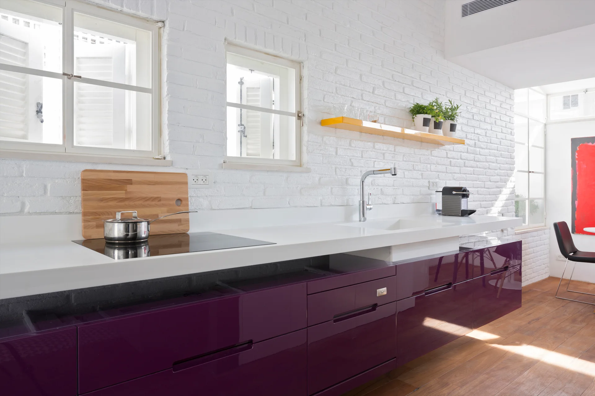 Glossy purple base cabinets and a white countertop sit against a white painted brick wall with two windows in a modern kitchen.