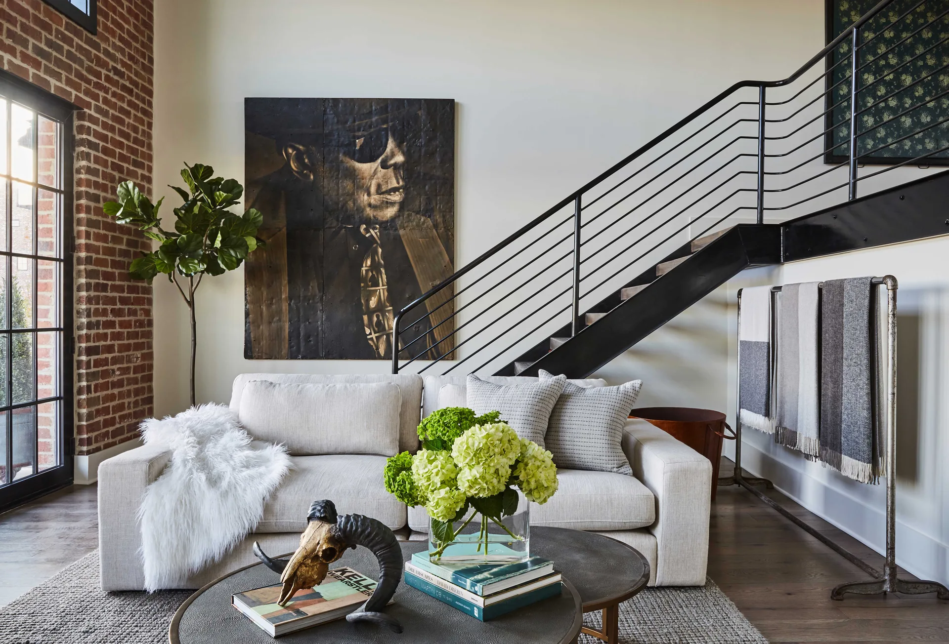 A cream sofa sits in front of a black metal staircase and a large portrait artwork, beside an exposed brick wall and industrial blanket rack.