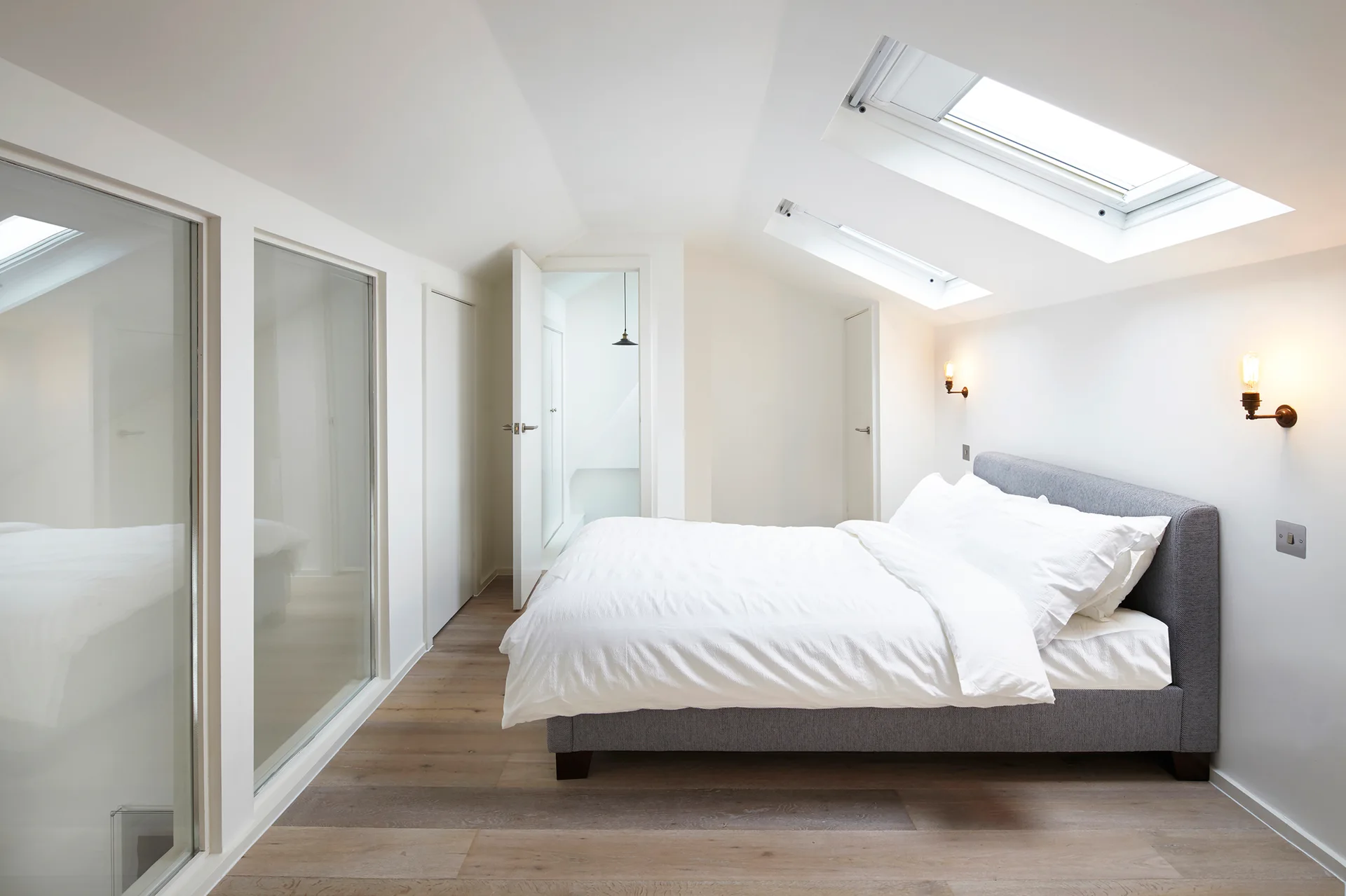 A modern attic bedroom features a grey upholstered bed under skylights, white walls, light wood flooring, and a glass partition.
