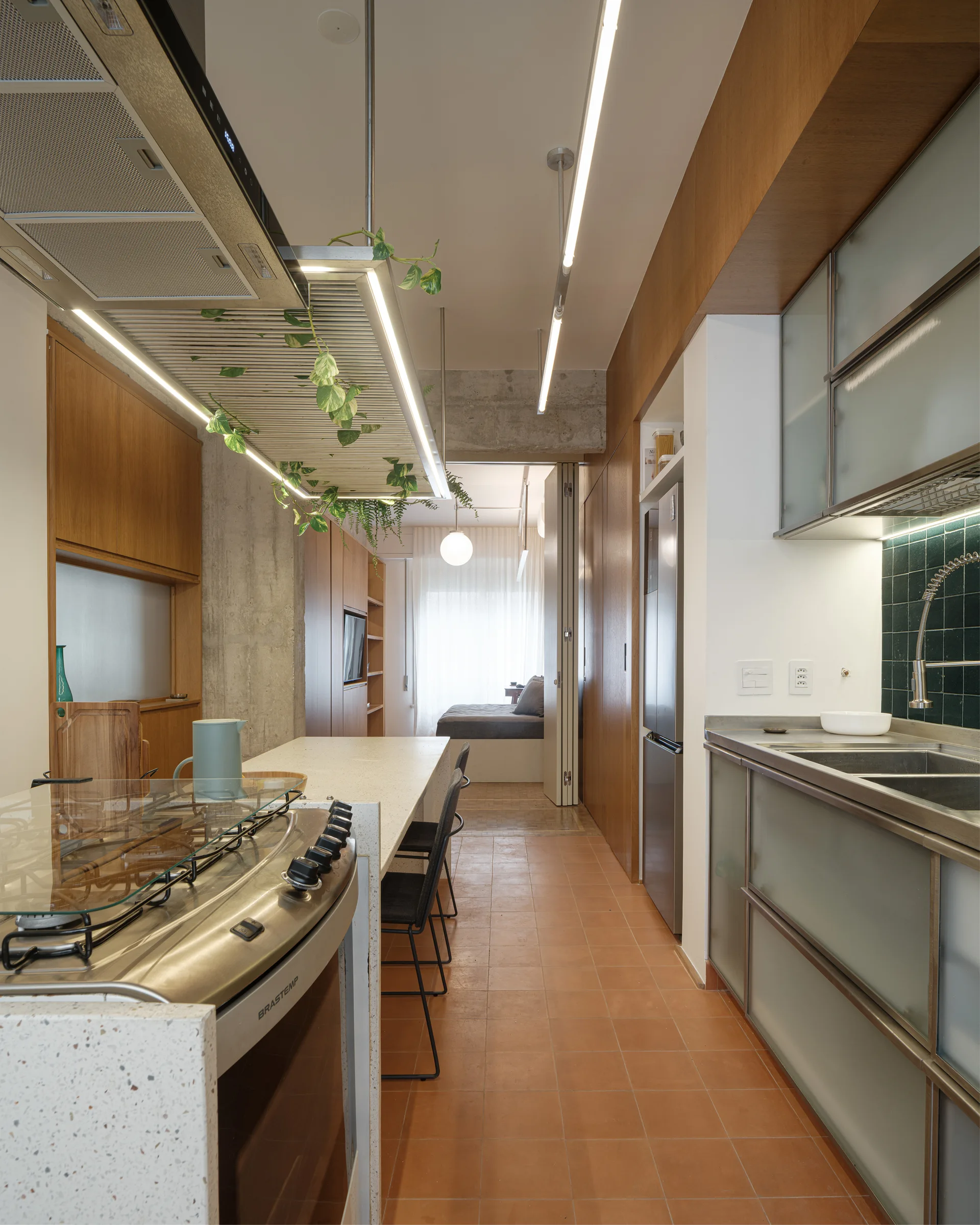A narrow modern kitchen features terracotta tile flooring, wood-toned cabinetry, a terrazzo-like island with a built-in stove, and a suspended plant rack.