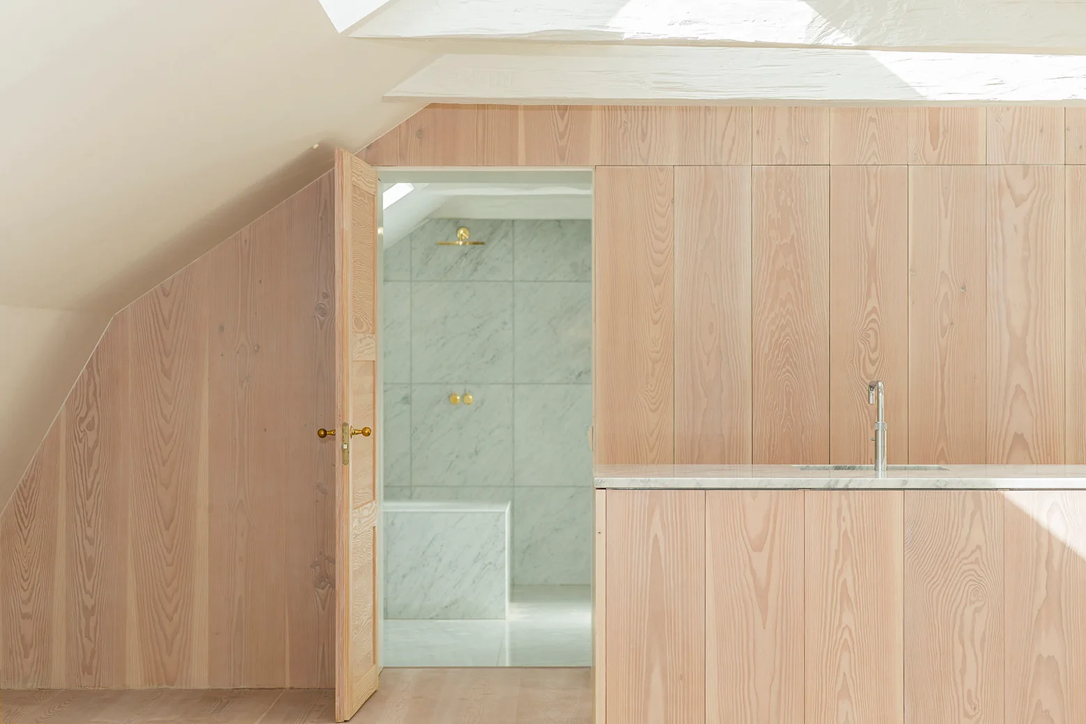 An open wood door in a pale wood-paneled attic space reveals a bright bathroom finished in veined white marble tile.