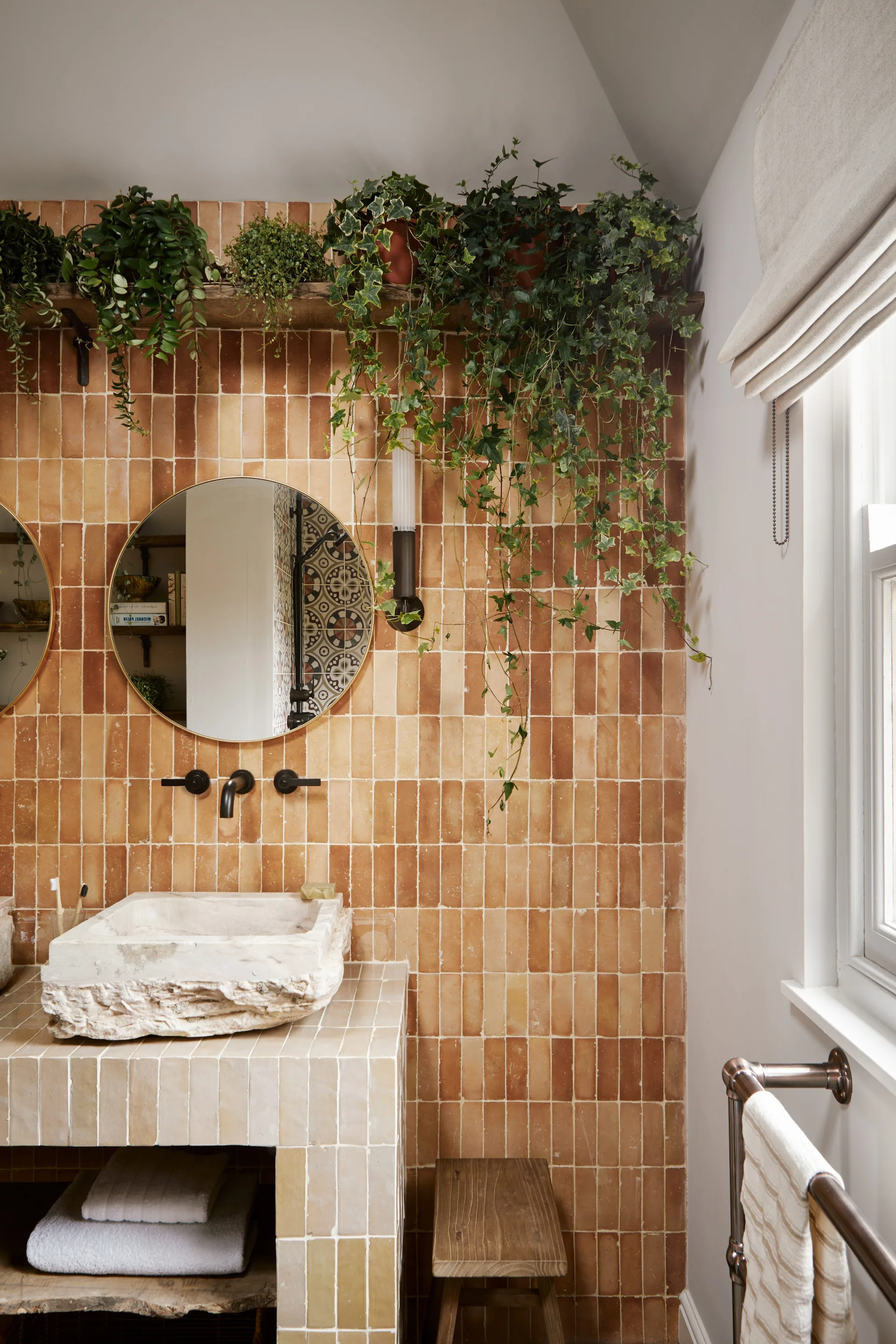 A bathroom featuring a raw stone vessel sink on a tiled vanity, set against a vertical terracotta tiled wall with cascading green plants on a shelf.