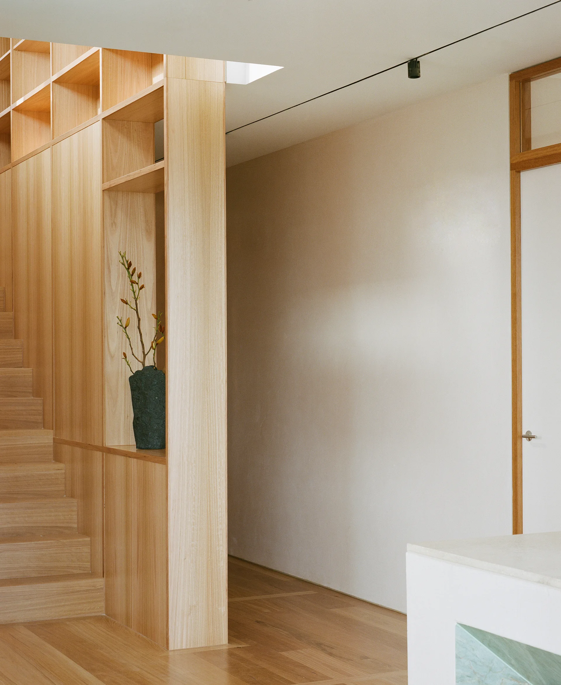 A minimalist light wood staircase features integrated shelving, wood paneling, and an adjacent white wall with wooden flooring.