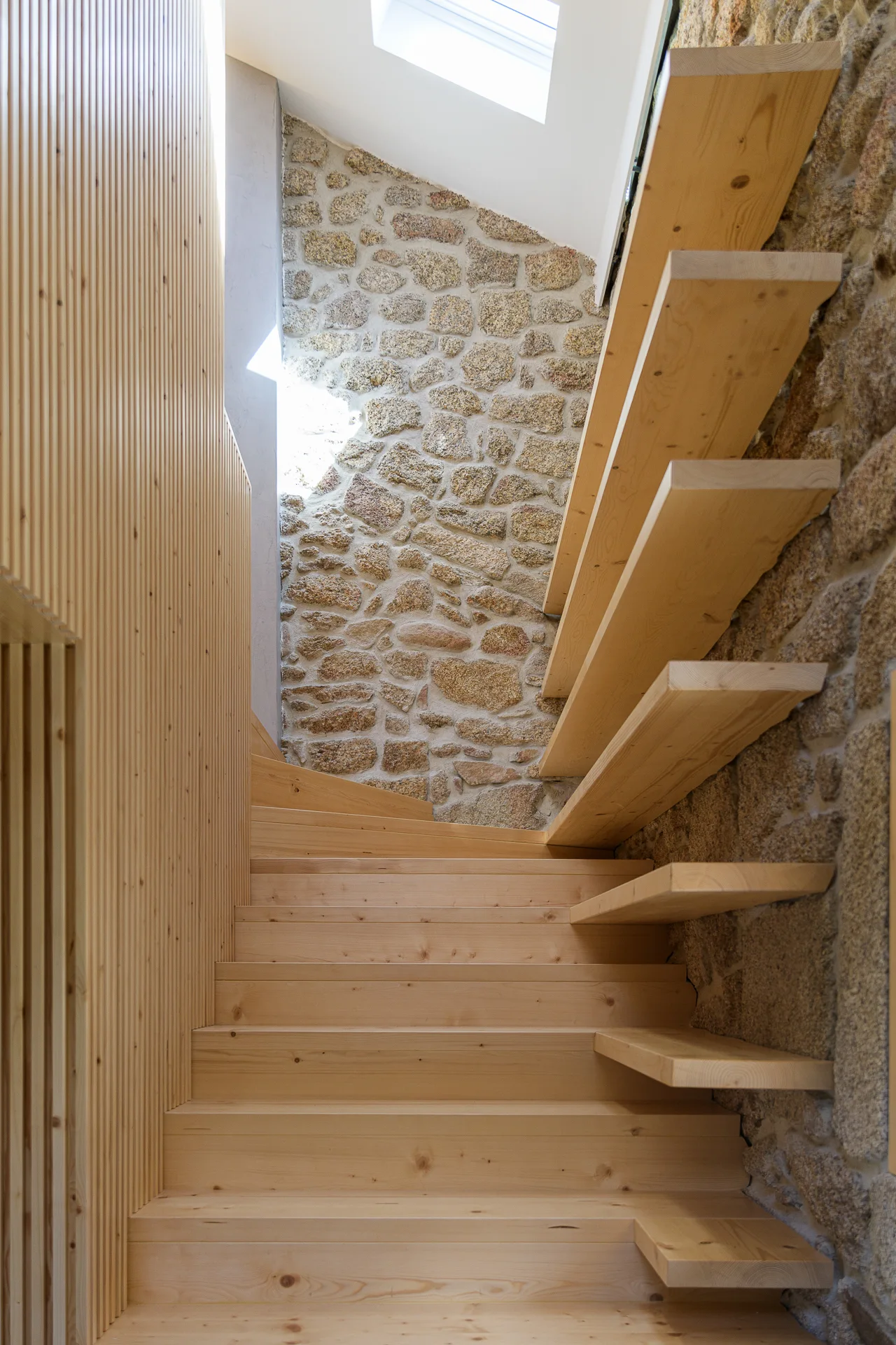 A natural wood staircase with floating treads ascends past an exposed stone wall and a vertical wood slatted panel under a skylight.