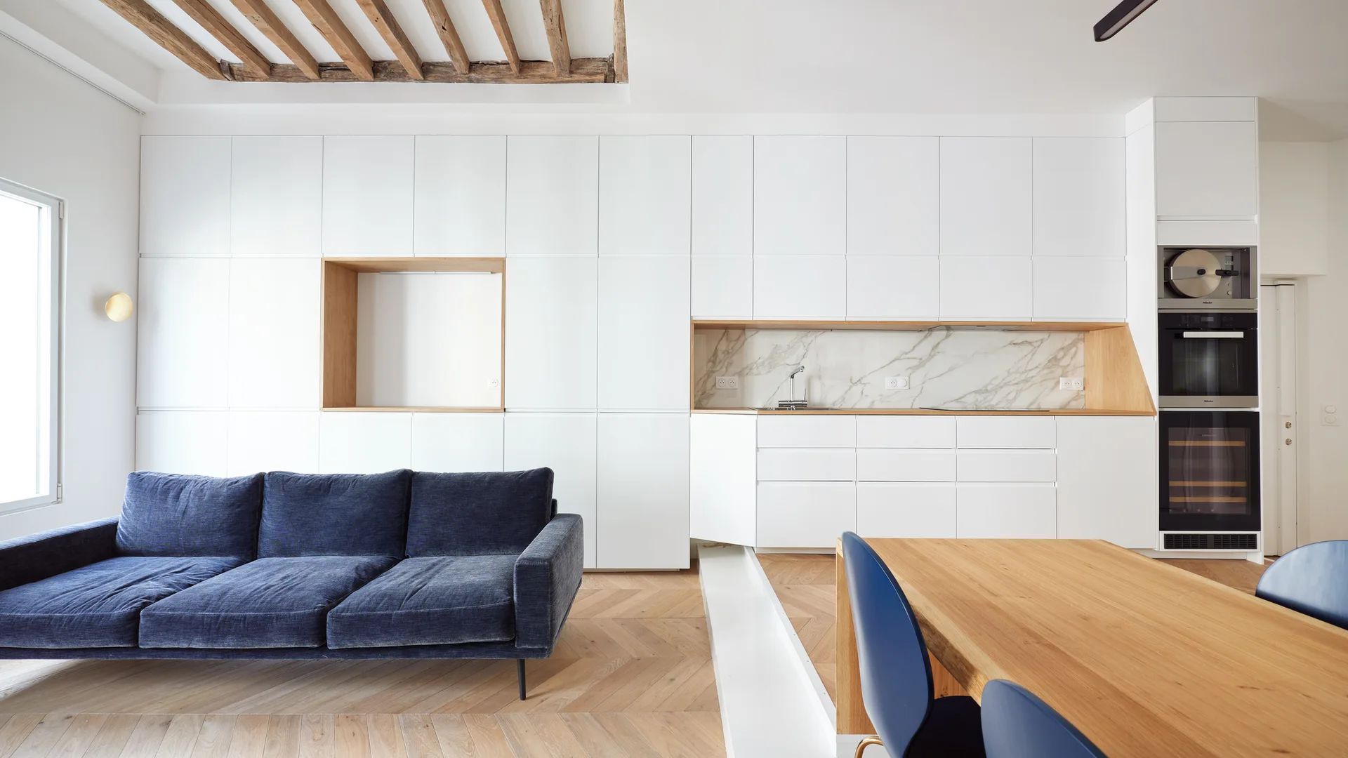A modern open-concept space featuring a deep blue sofa and dining area set against a seamless white storage wall with integrated kitchen features.