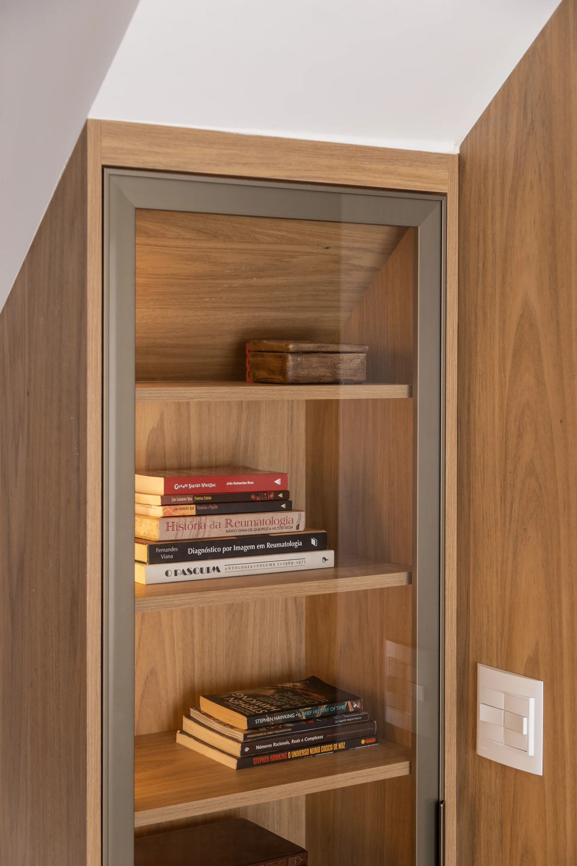 A built-in wood display cabinet under a sloped ceiling features warm shelf lighting, glass doors, and books.