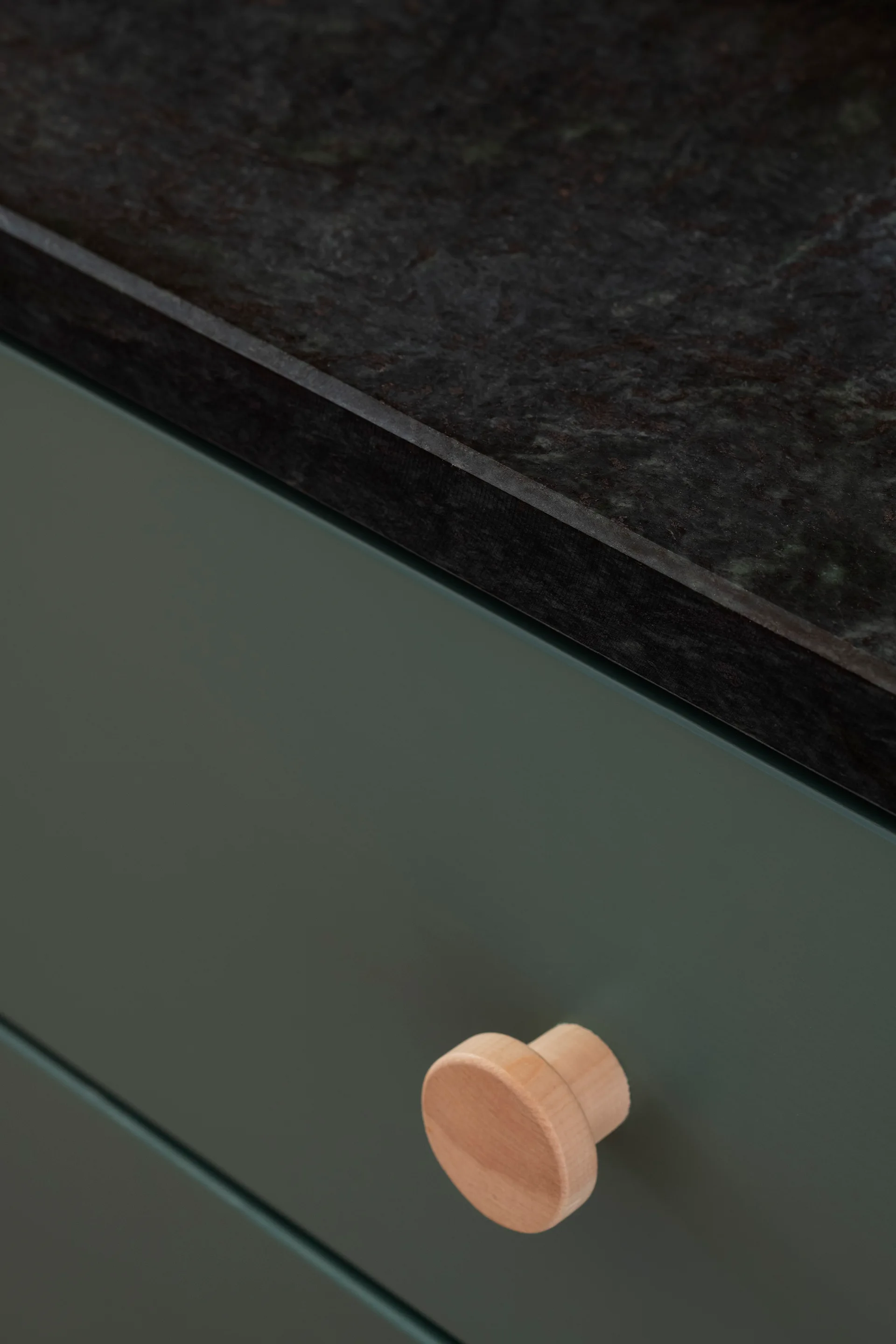 A close-up of a matte green cabinet drawer with a round light wood knob below a dark textured stone countertop.