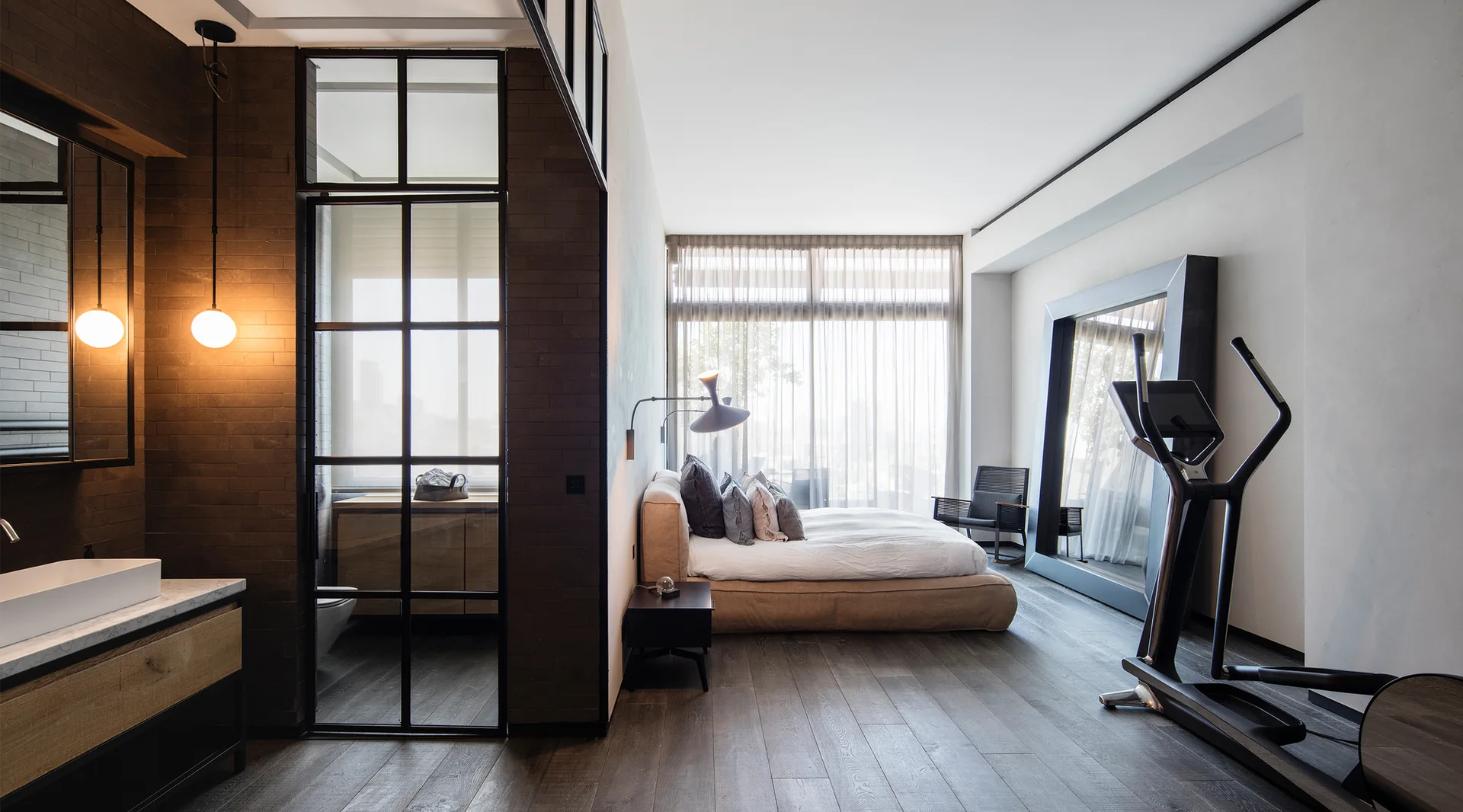 A modern bedroom features a low-profile platform bed, a steel-framed glass partition wall leading to an adjacent bathroom vanity, and an exercise machine.