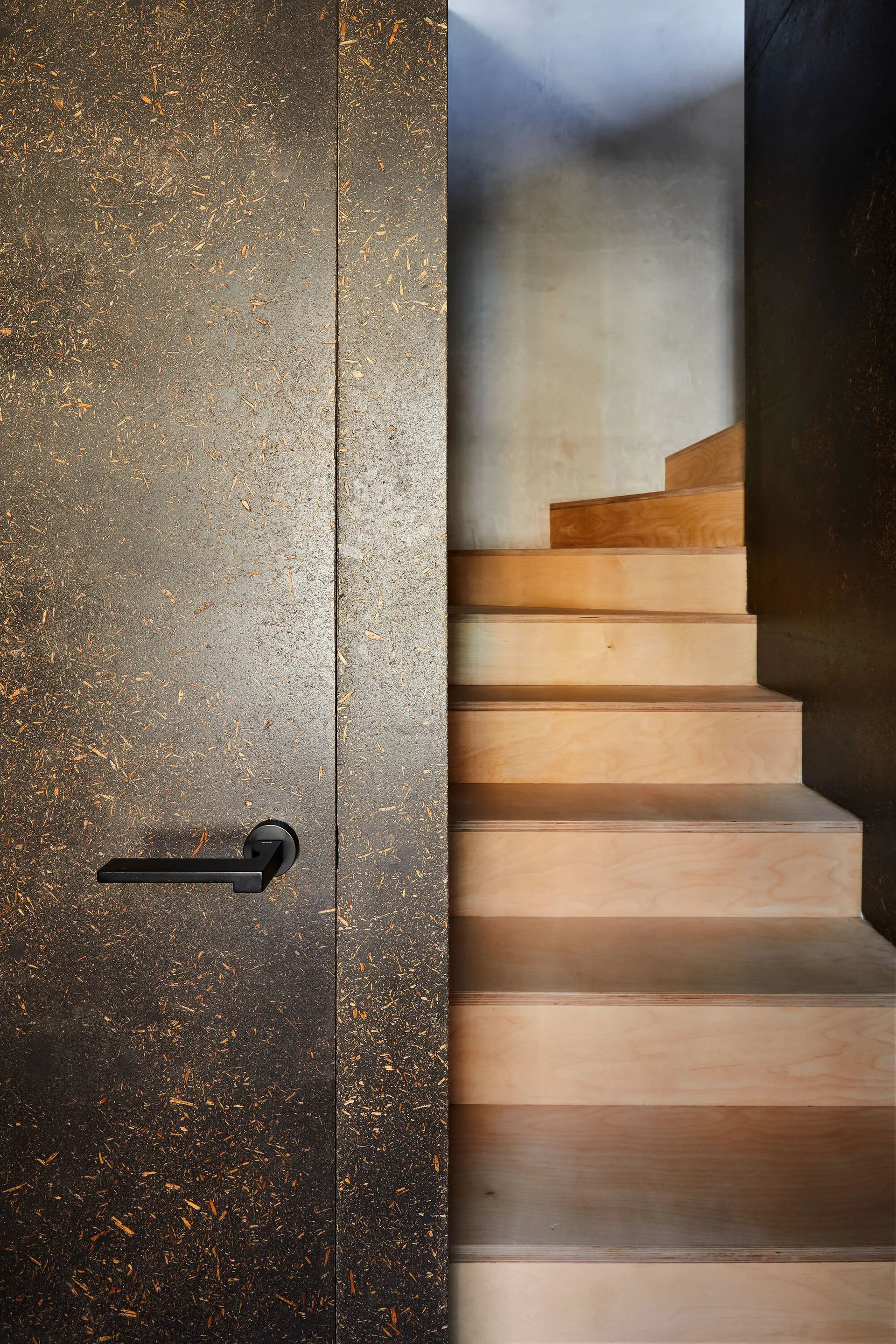 A dark speckled composite door with a black lever handle stands next to a modern light wood staircase.