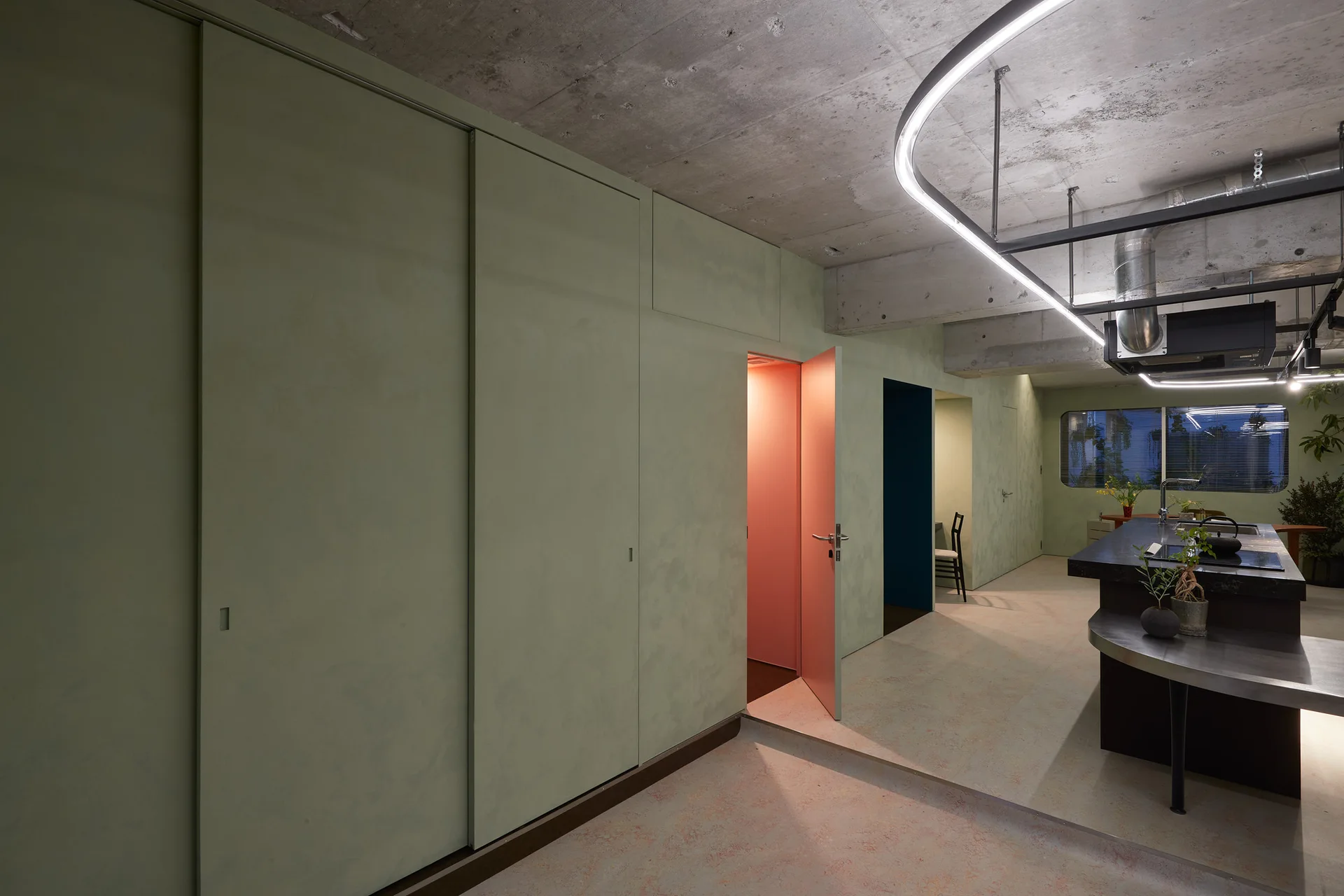 A minimalist industrial kitchen featuring sage green sliding wall panels, an exposed concrete ceiling, a black island, and a partially open pink door.