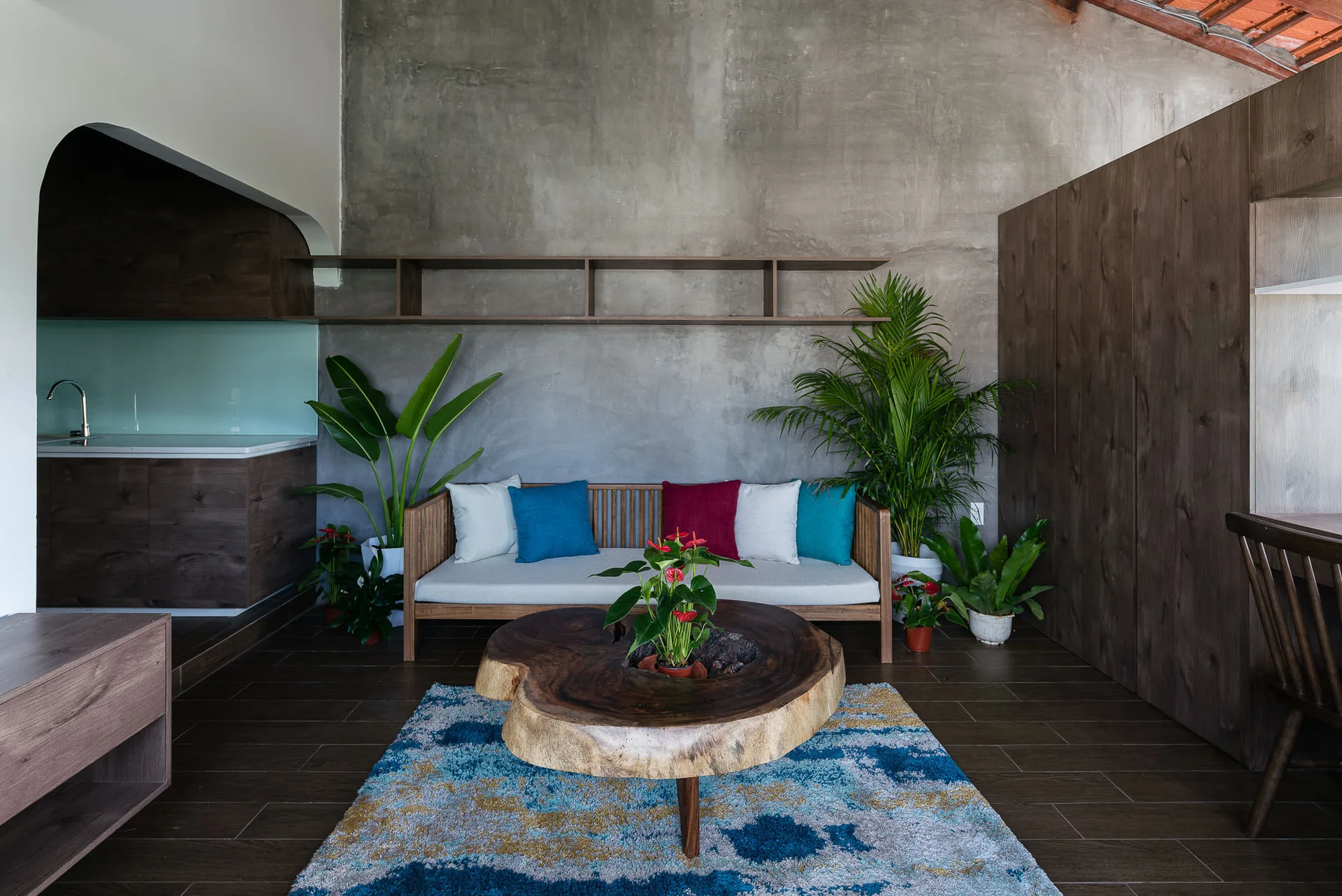 A wooden sofa with colorful throw pillows and a live-edge coffee table sit on a patterned blue rug against a grey concrete wall.