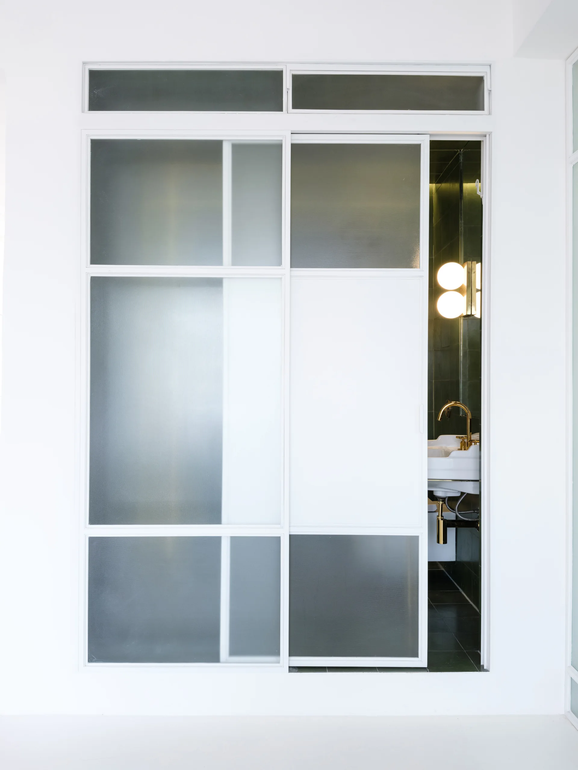 A frosted glass sliding door in a white wall partially opens to reveal a dark green tiled bathroom with a gold faucet and glowing sconce.