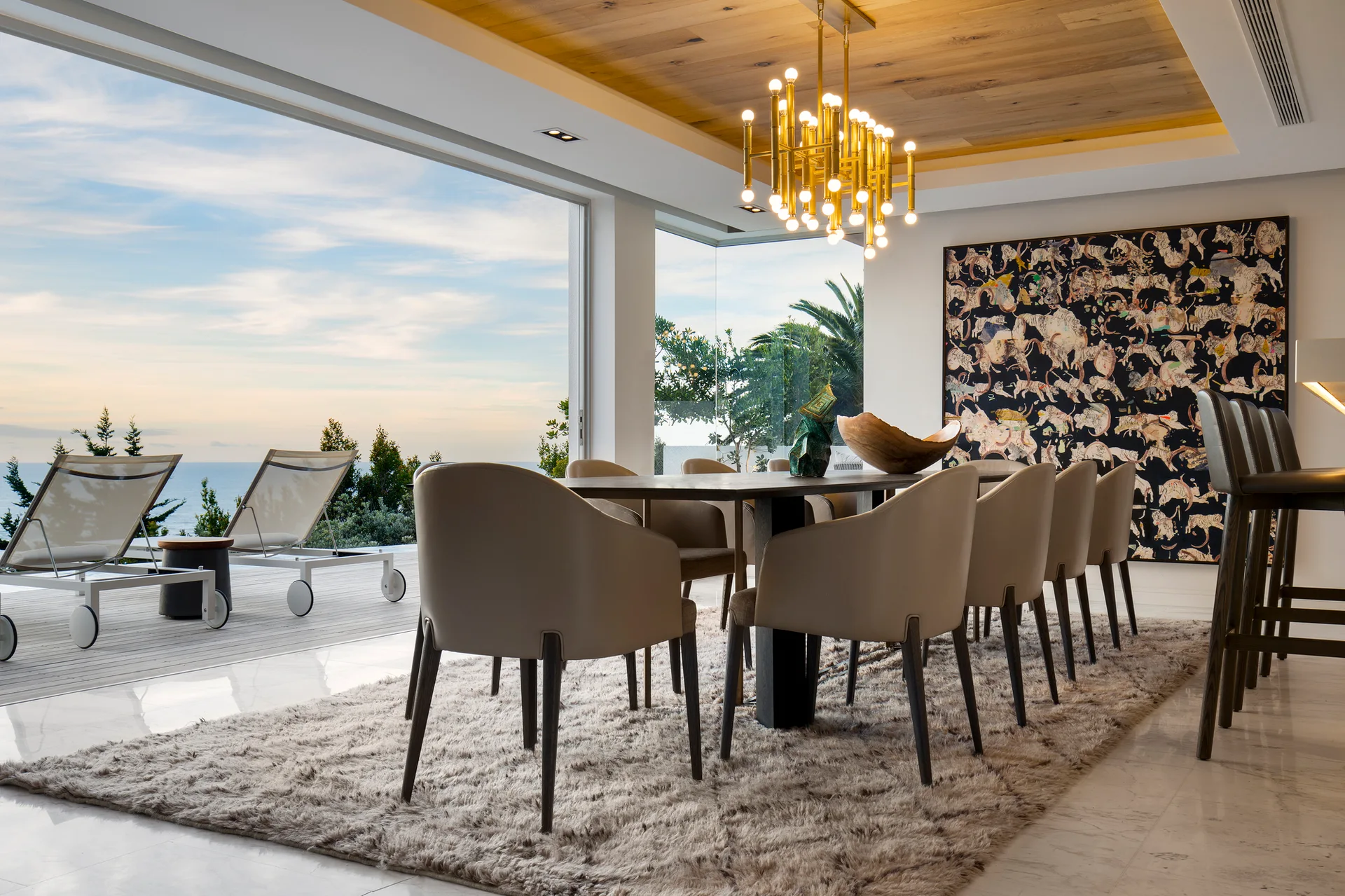 A modern dining room features a long table with beige chairs, a gold chandelier, and an inset wood ceiling panel overlooking the sea.