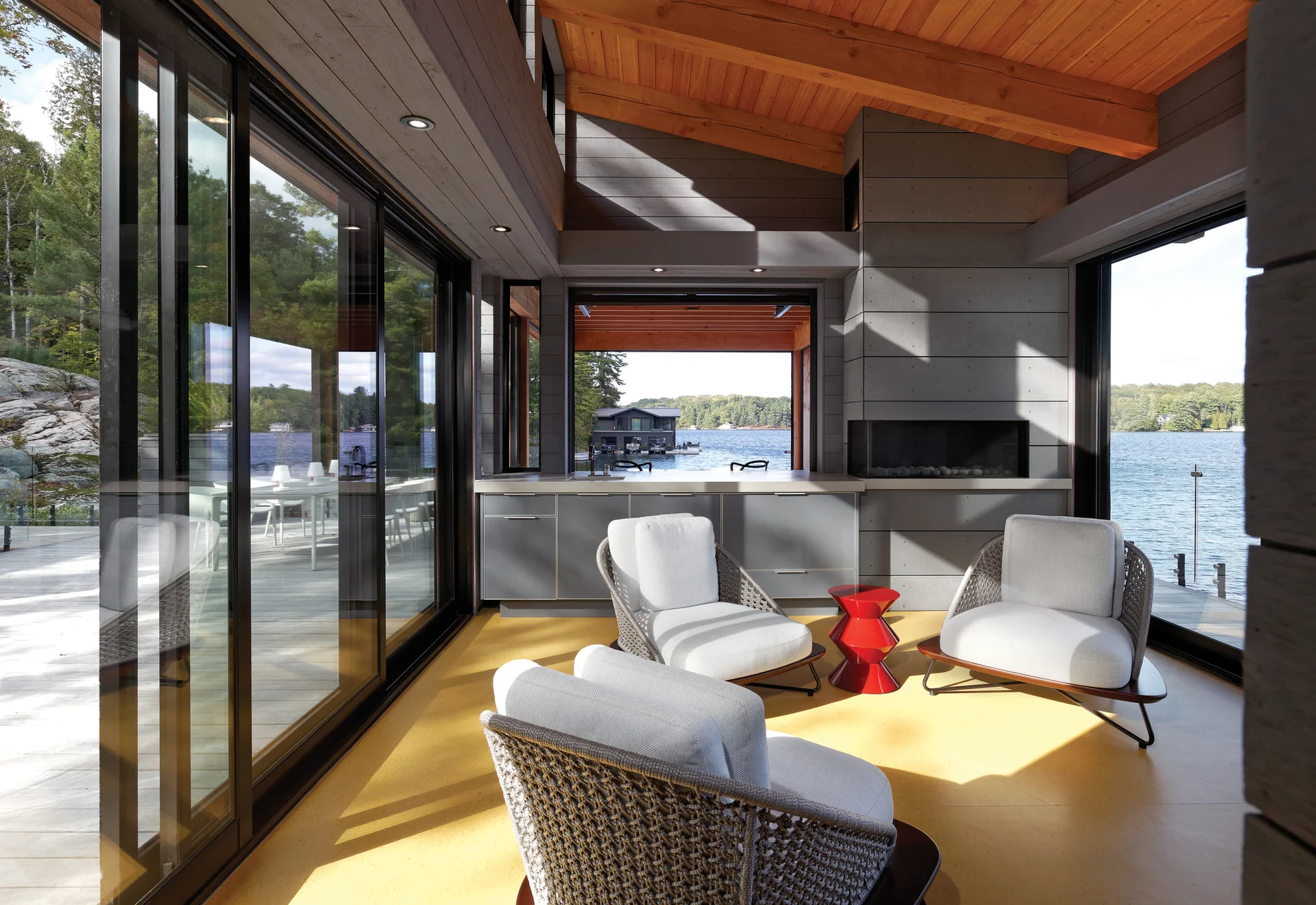 A lakeside lounge features three white chairs on a bright yellow floor with grey cabinetry and a natural wood plank ceiling.