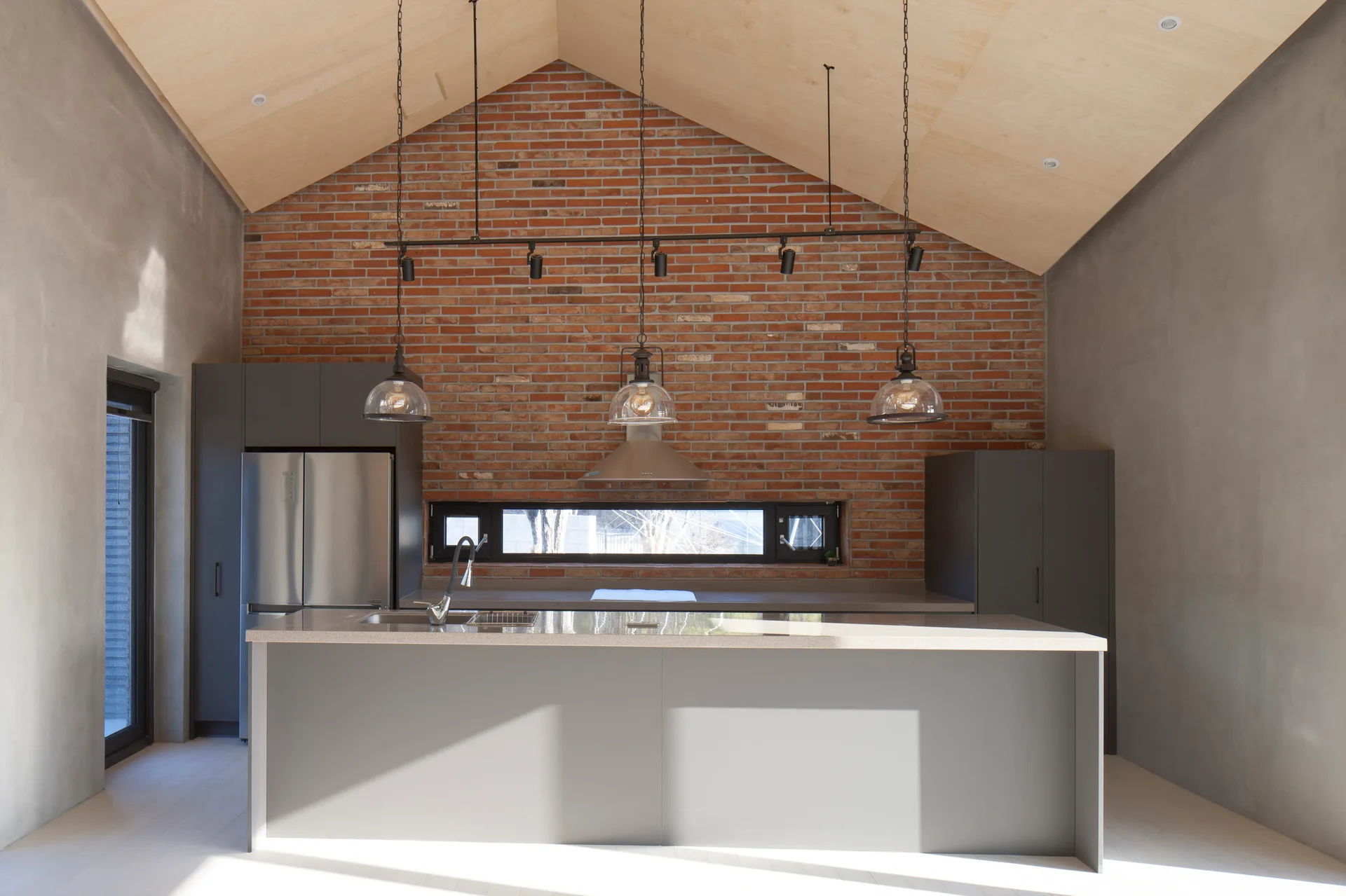 A modern kitchen featuring a gray island, an exposed brick back wall, and a vaulted plywood ceiling with three glass pendant lights hanging above.