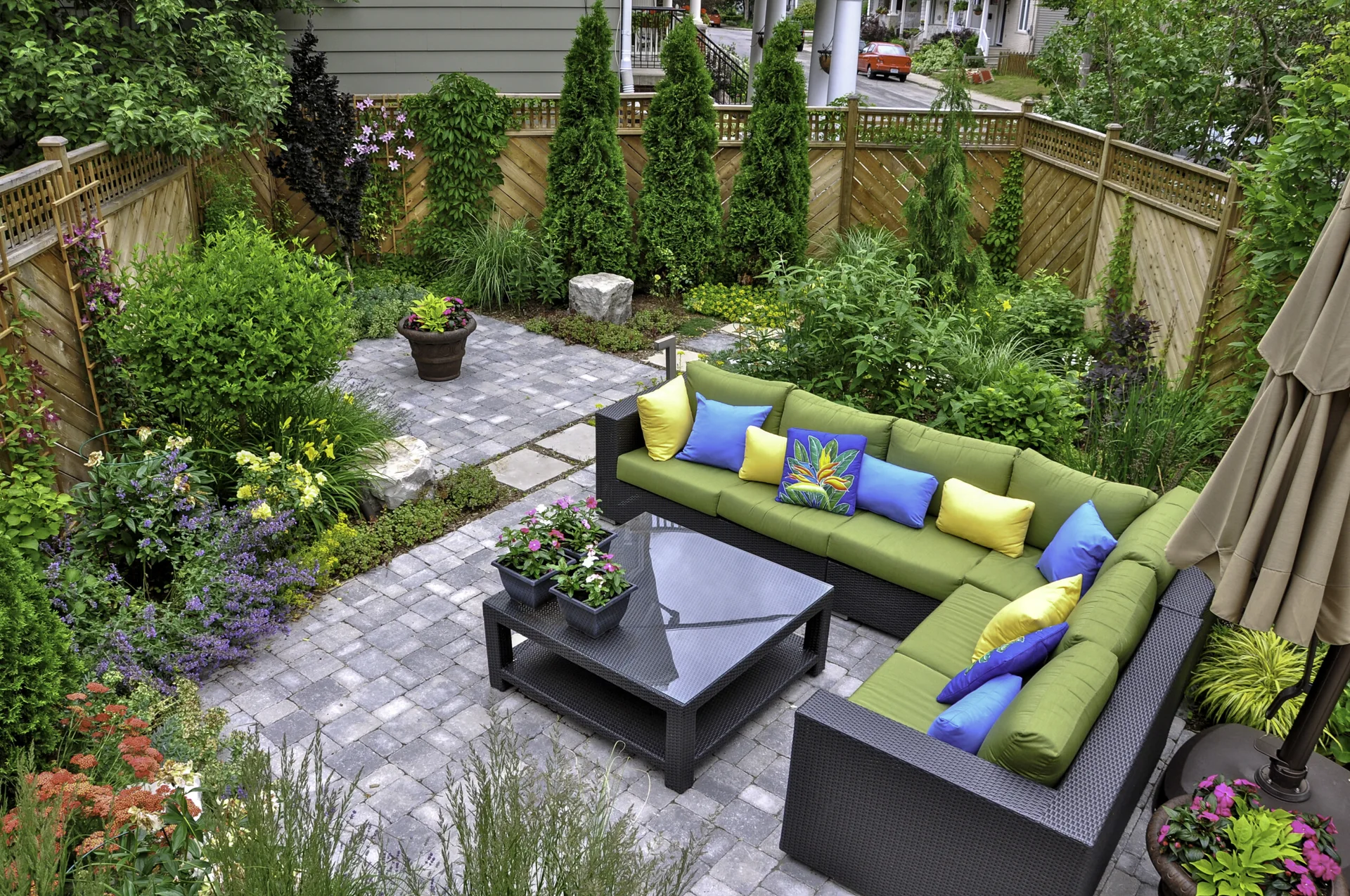 A backyard stone paver patio features a dark wicker-style sectional sofa with green cushions, colorful pillows, and a glass-topped coffee table.