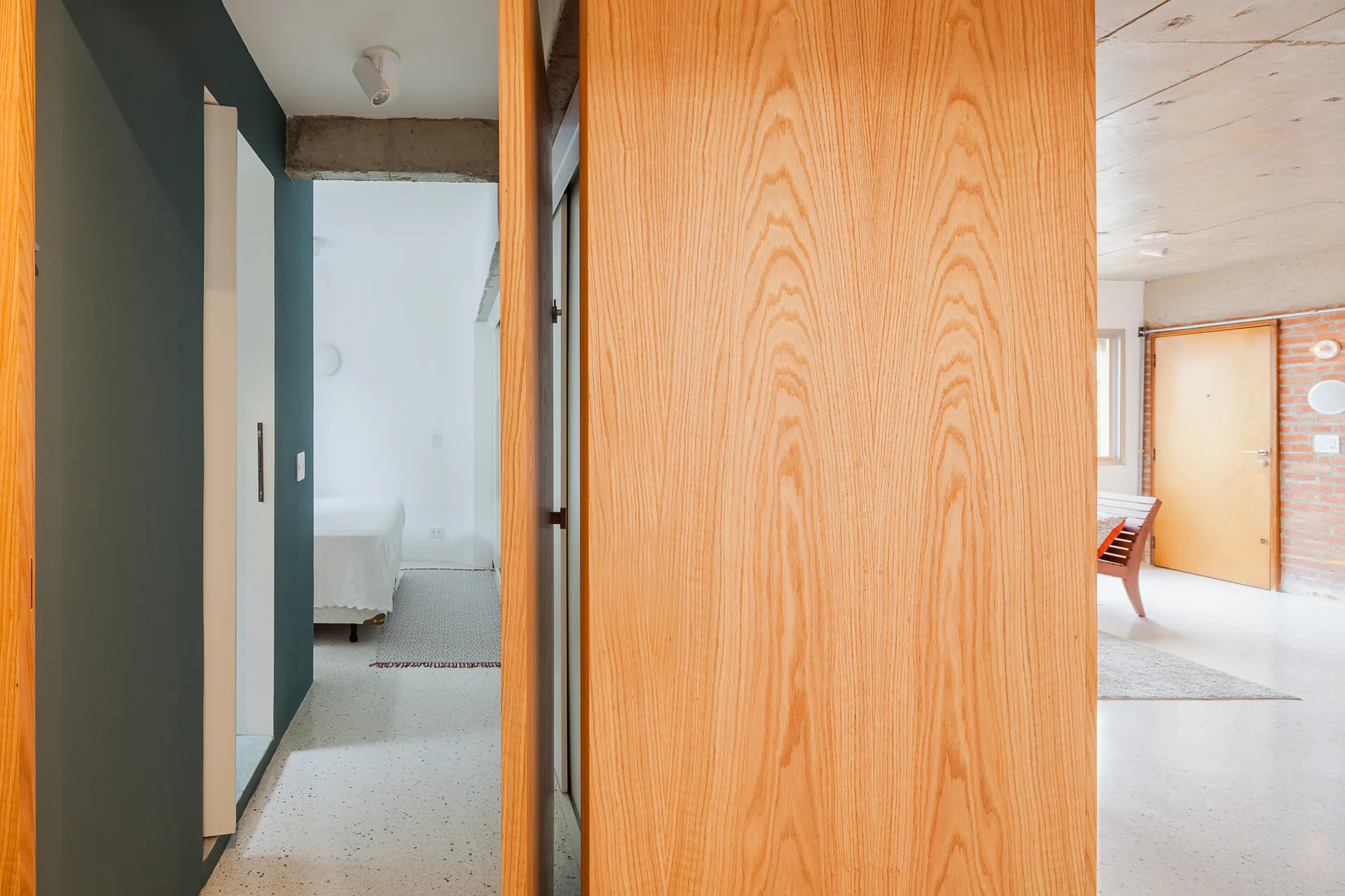 A view down a speckled hallway with a dark blue-grey wall and a large wood grain door frame looking into a bedroom and adjacent living space.