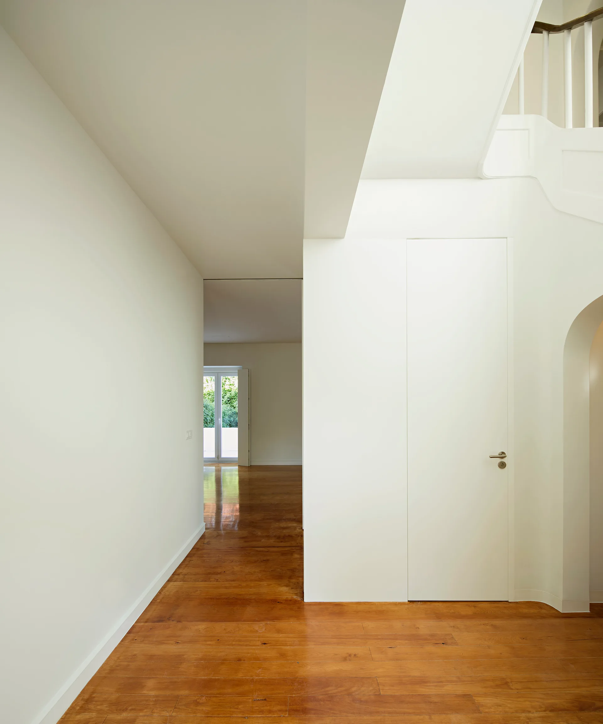 A minimalist hallway features bright white walls, a flush door, warm hardwood plank floors, and a white staircase leading upstairs.