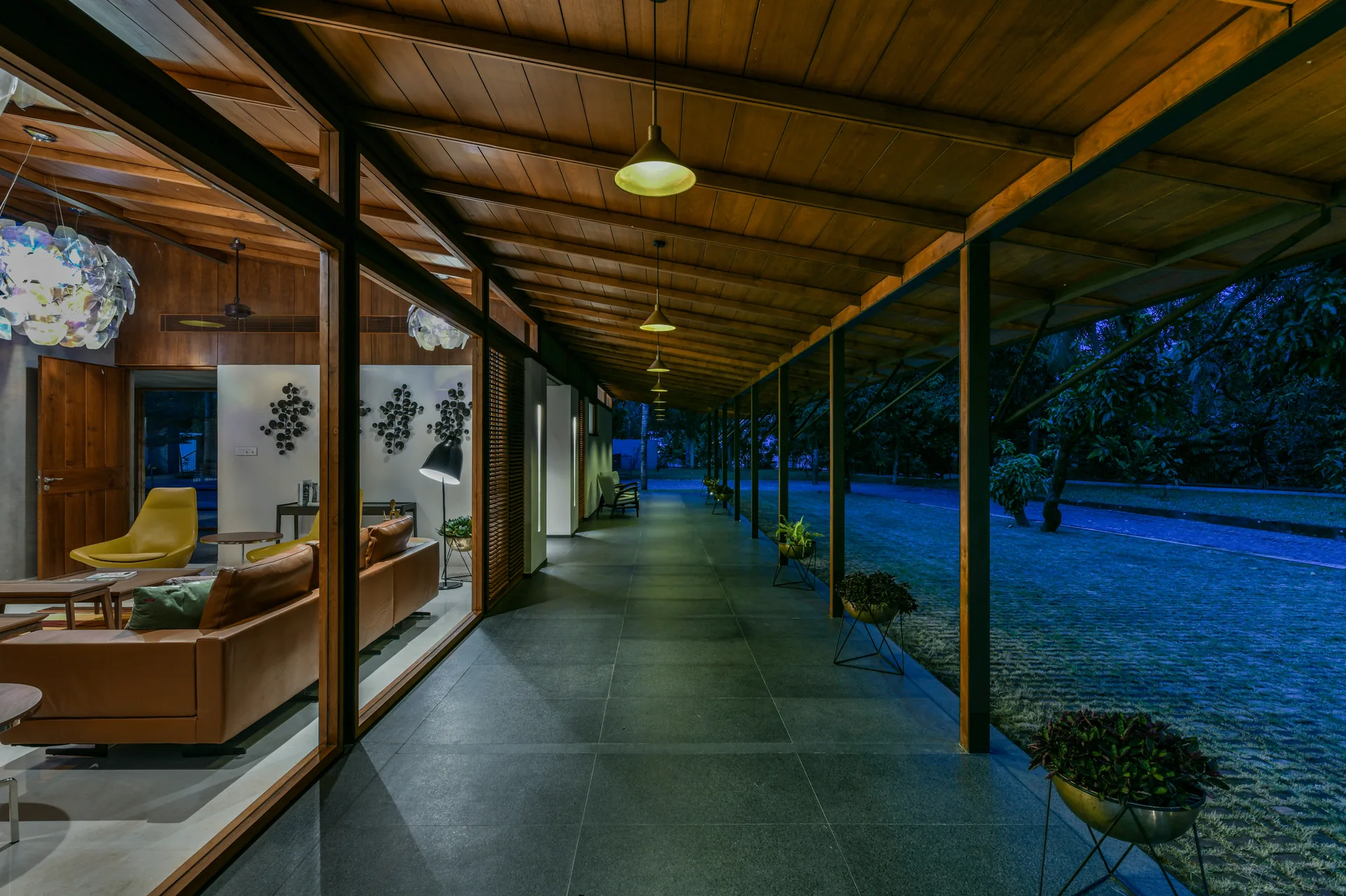 A covered patio walkway with a wood ceiling and dark stone tiles overlooks a lawn at night next to a glass-walled lounge.