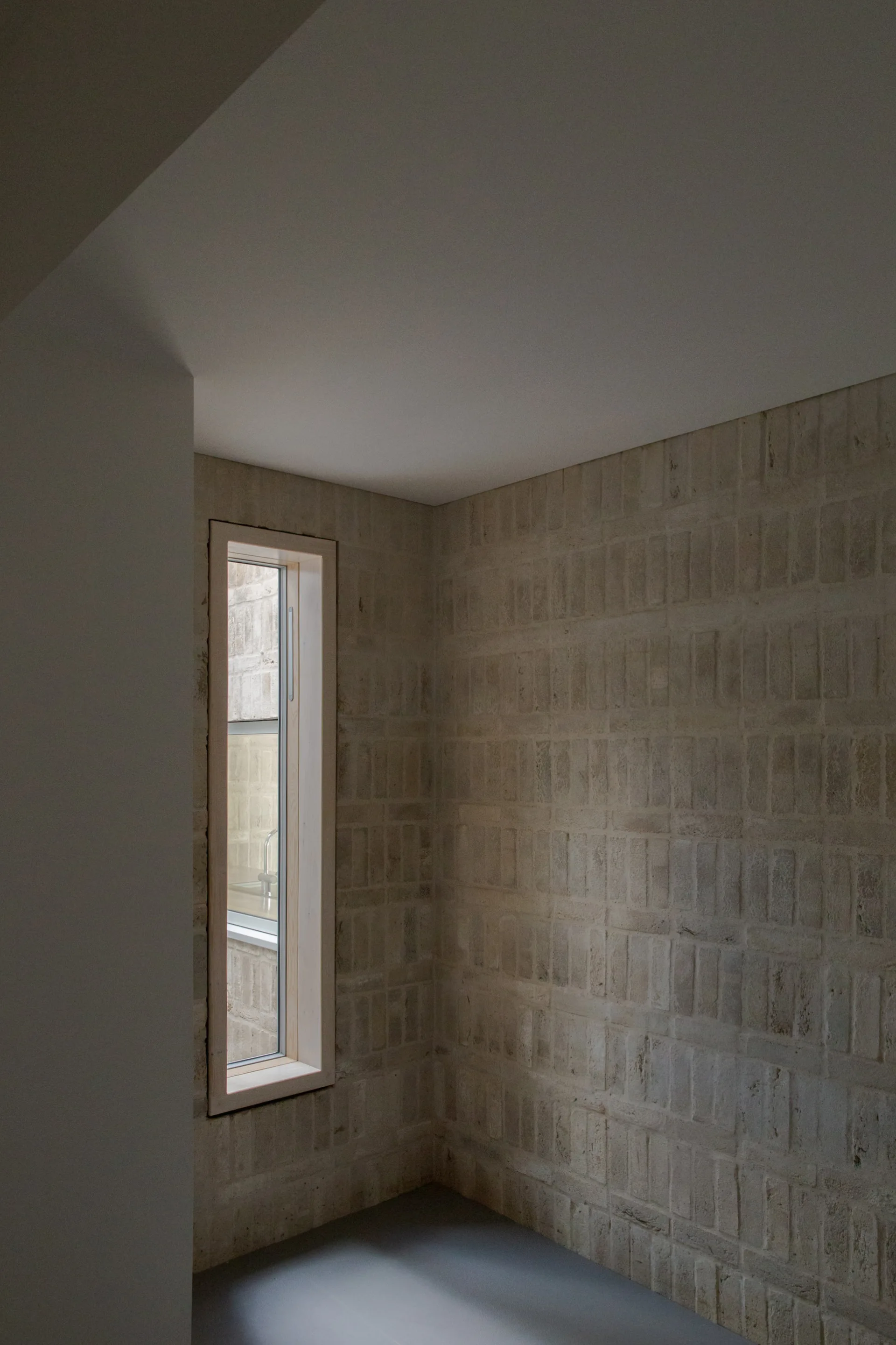 An interior corner features exposed tan masonry brick walls, a smooth grey floor, and a tall narrow window framed in light wood.