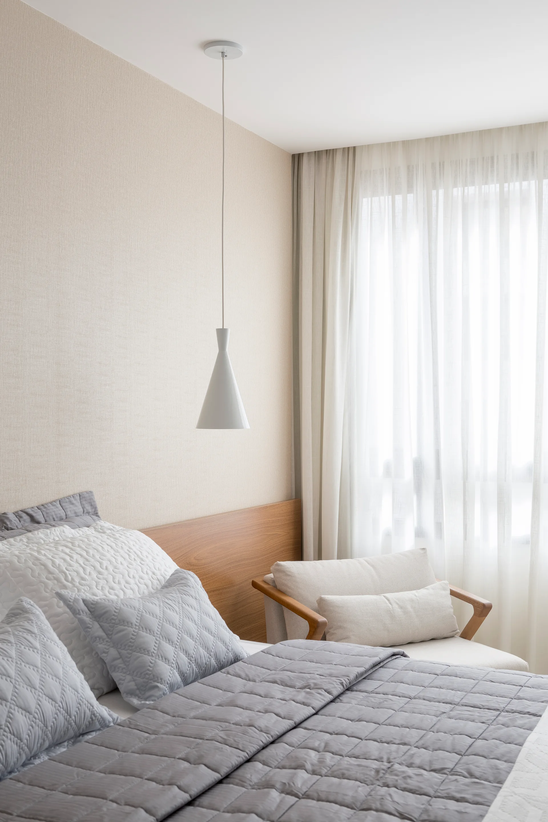 A minimalist bedroom featuring a low wooden headboard, grey quilted bedding, a white pendant light, and an armchair by sheer curtains.