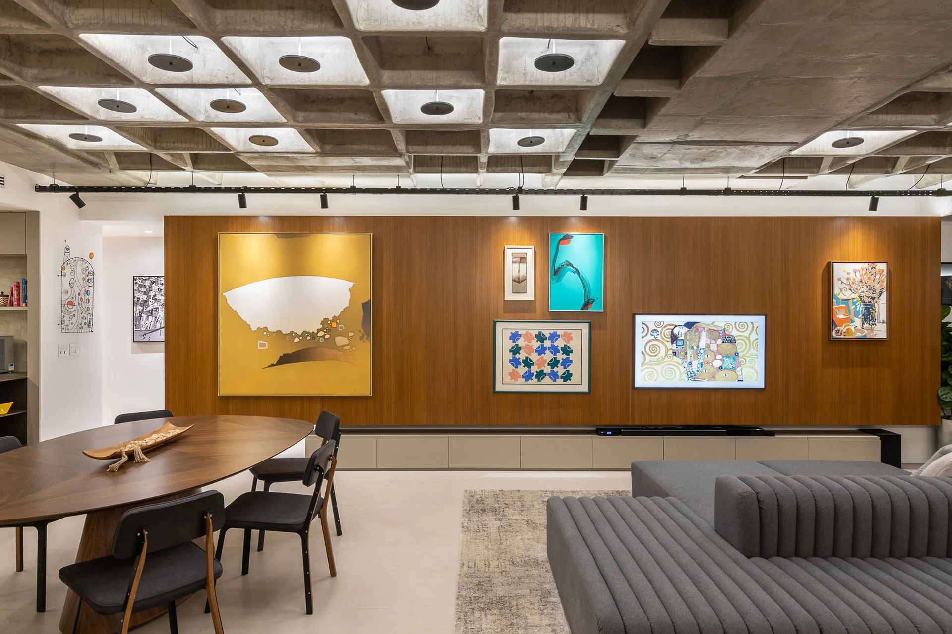 A modern living area features an industrial concrete waffle ceiling, a warm wood-paneled wall with framed art, a grey ribbed sofa, and a dining set.