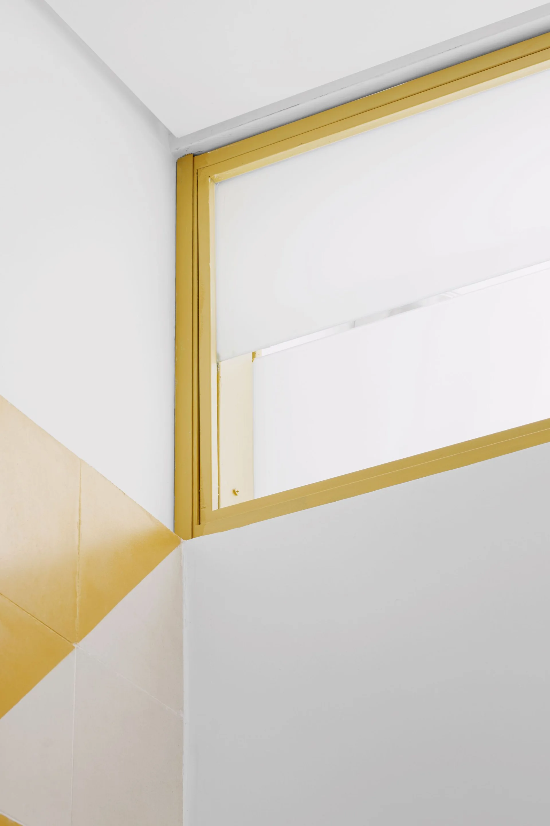 A close-up of a bright yellow painted window frame set against a white wall next to a geometric yellow and white tiled corner.