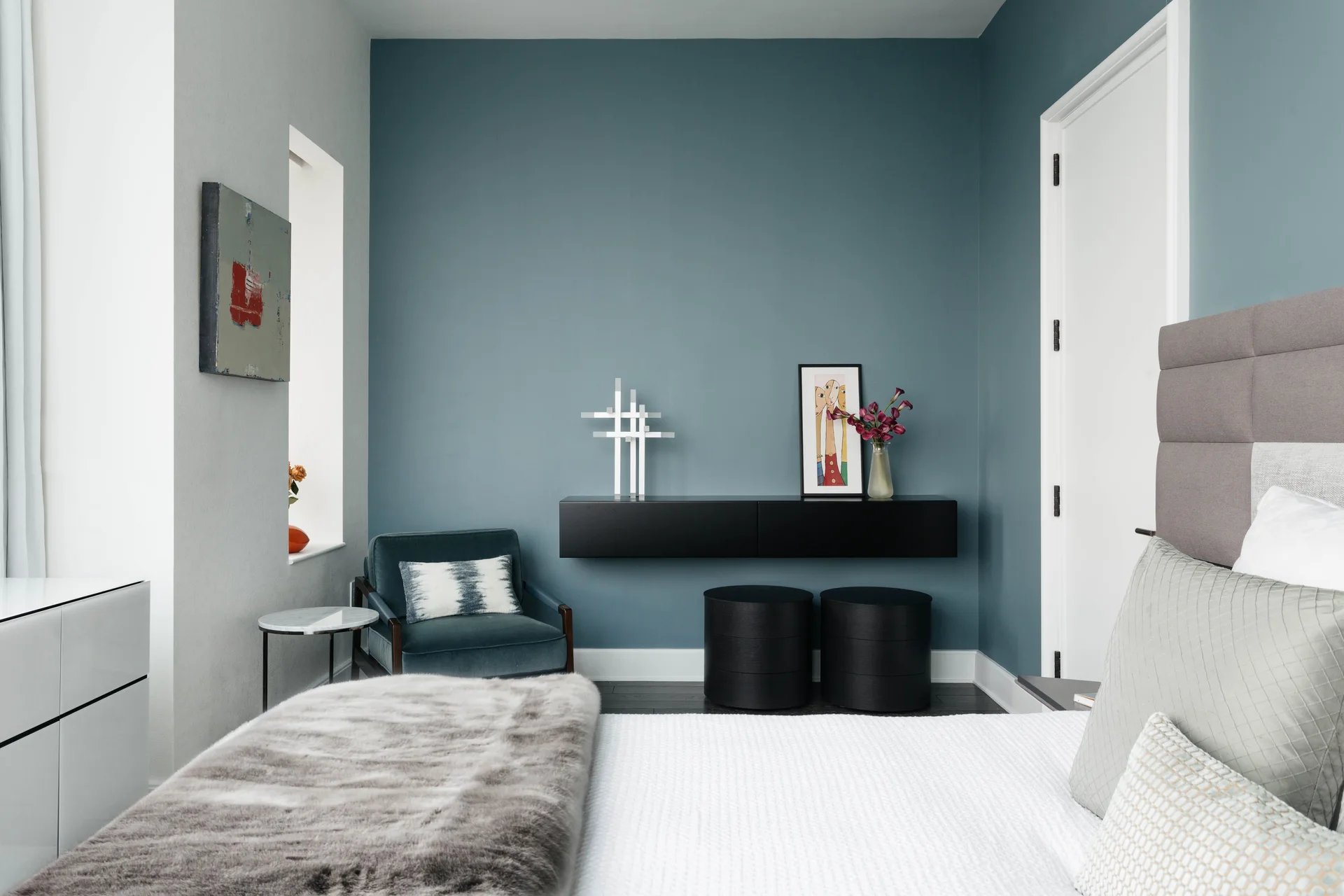 A modern bedroom featuring a dark blue accent wall with a wall-mounted black console, two cylindrical black stools, and a grey upholstered bed.