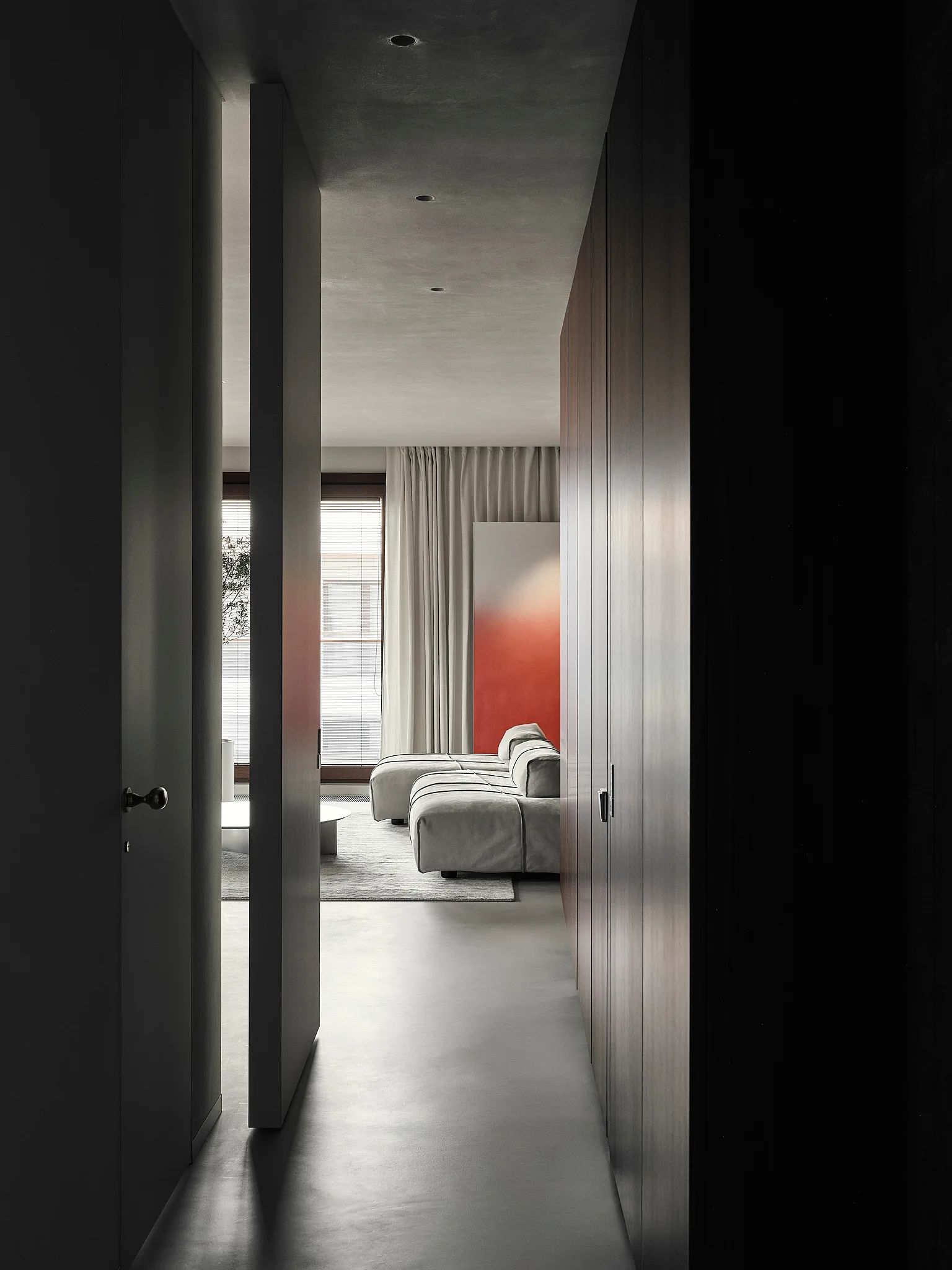 A view down a dark grey hallway with wood cabinetry toward a bright living room featuring a grey modular sofa and large orange artwork.