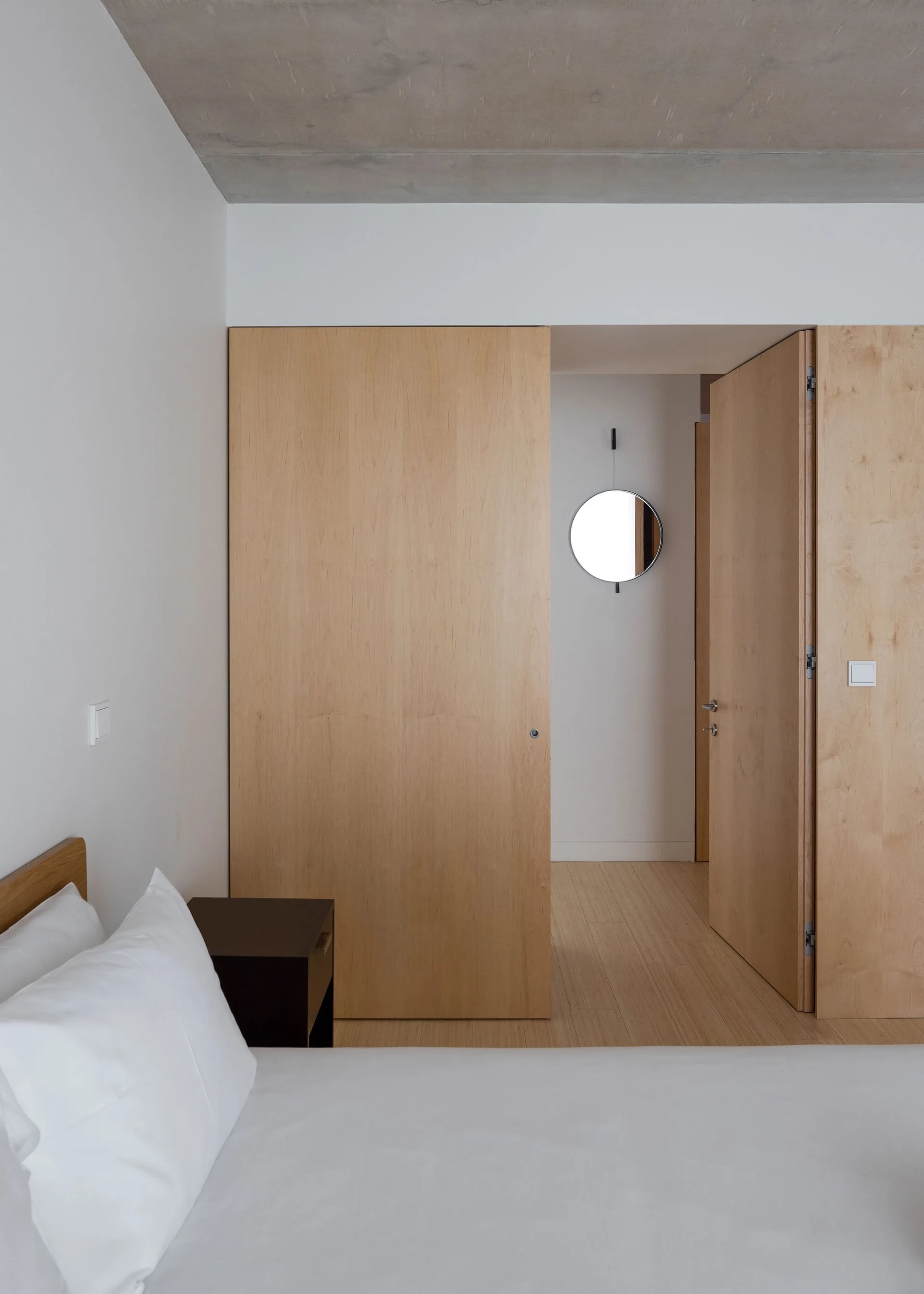 A minimalist bedroom features an exposed concrete ceiling, light wood sliding doors, and a dark brown nightstand next to white bedding.