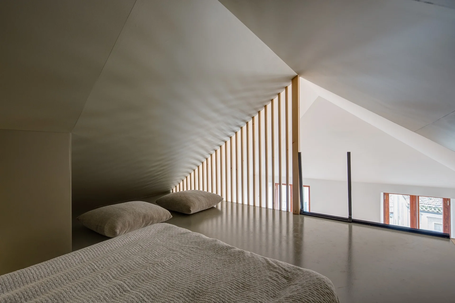 A minimalist attic loft bedroom features a low bed under a sloped ceiling, bordered by a vertical wooden slat railing that overlooks the lower floor.