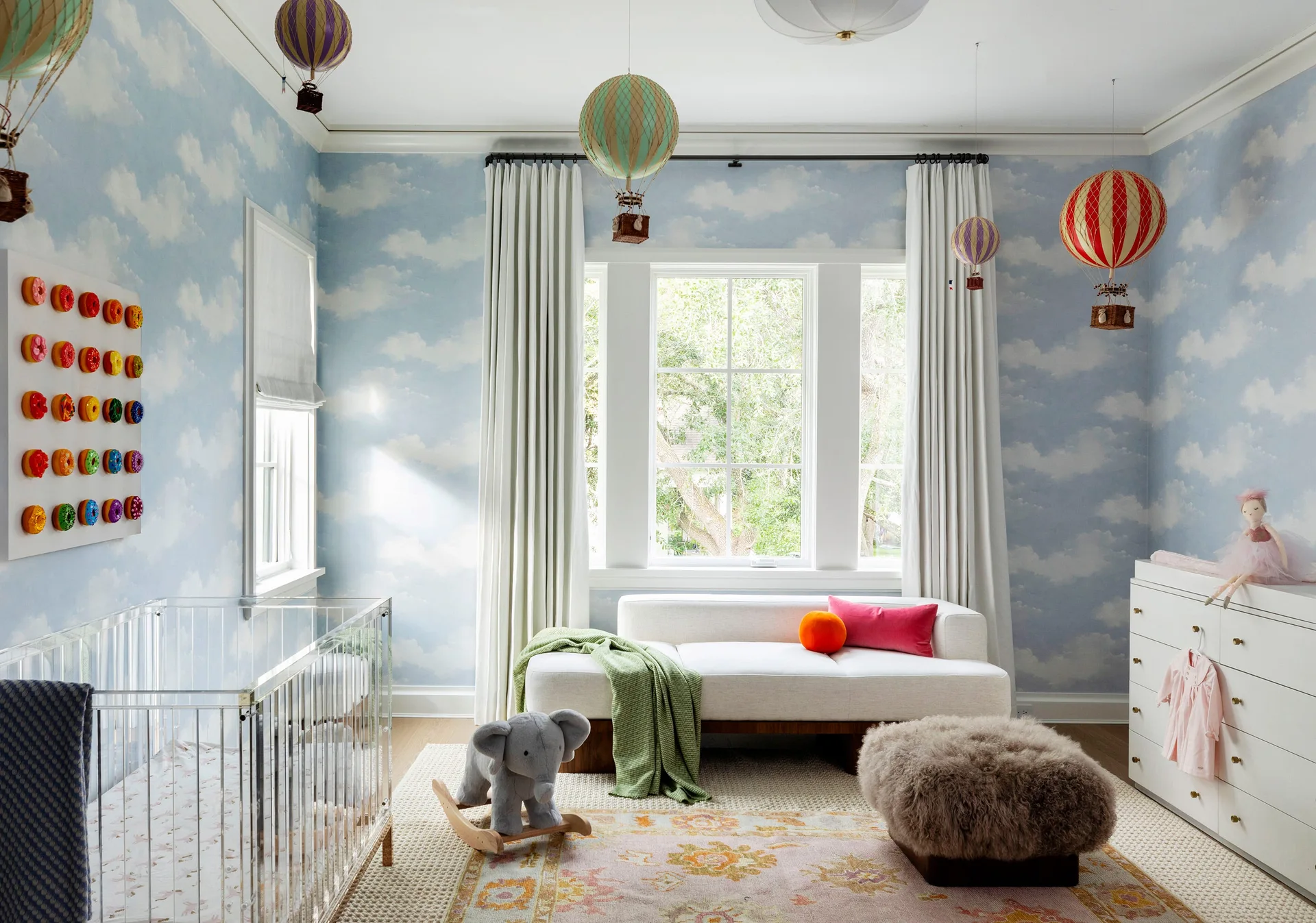 A whimsical nursery features sky blue cloud wallpaper, hanging hot air balloons, a clear acrylic crib, and a white daybed.