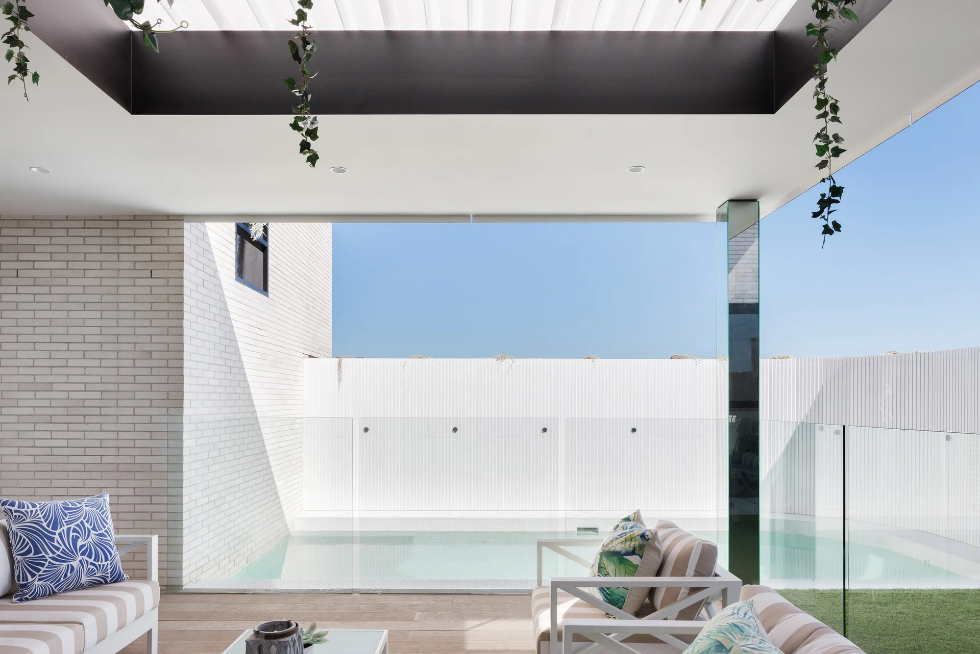 A modern covered patio lounge with white metal seating features a glass railing overlooking a small swimming pool next to a white brick wall.