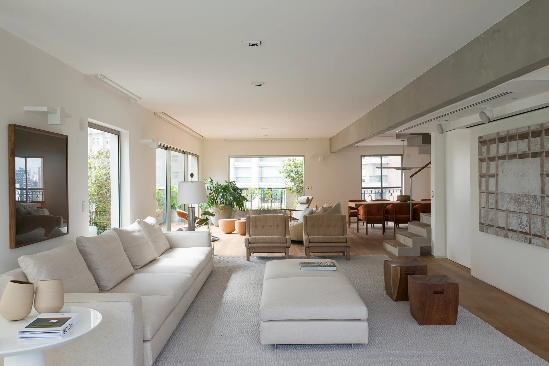 A spacious open-plan living room features a long cream sofa, two tan armchairs, wood cube stools, and an exposed concrete ceiling beam.