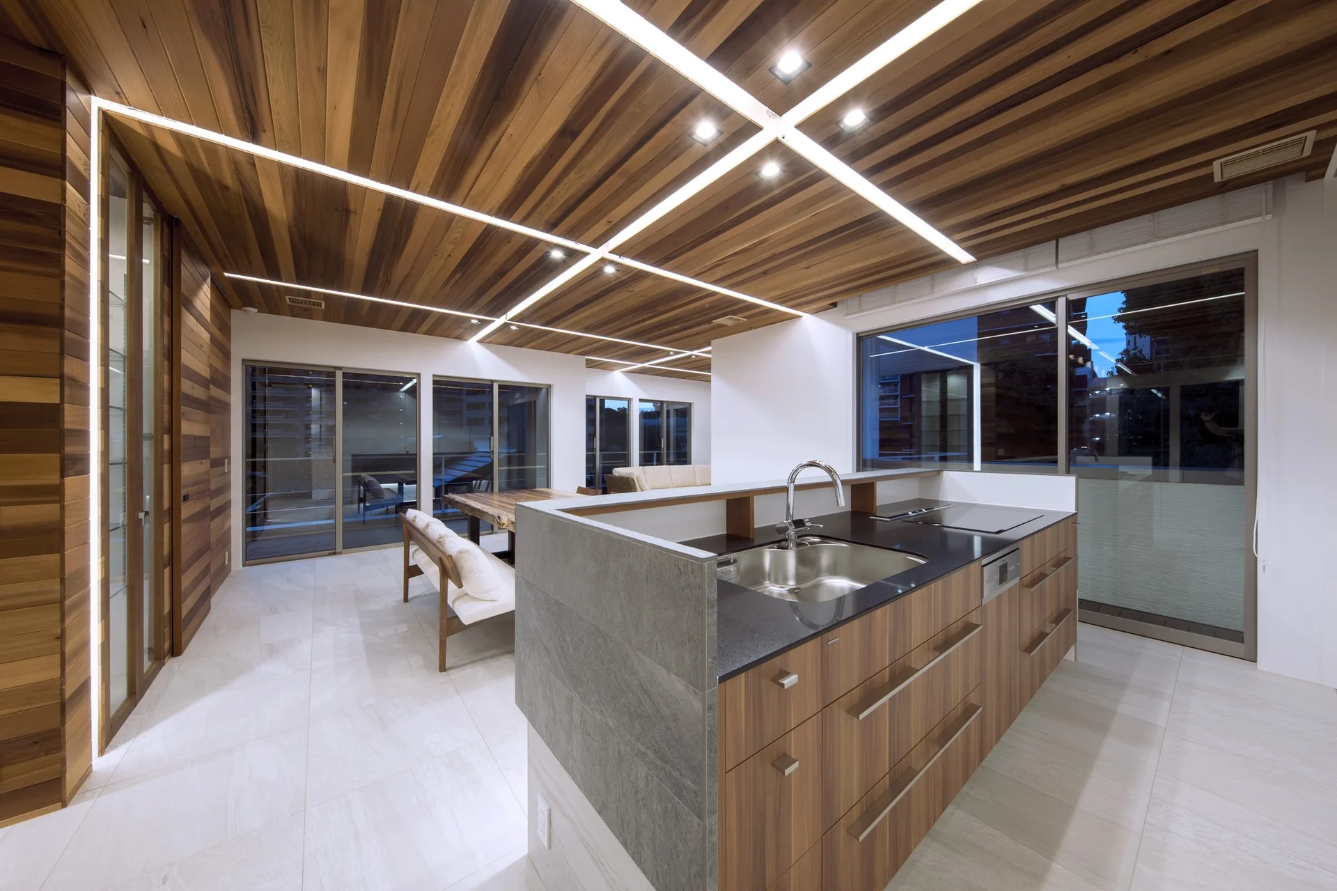 A modern kitchen features a central island with a wood finish, a dark countertop, and a wood plank ceiling with integrated linear light strips.