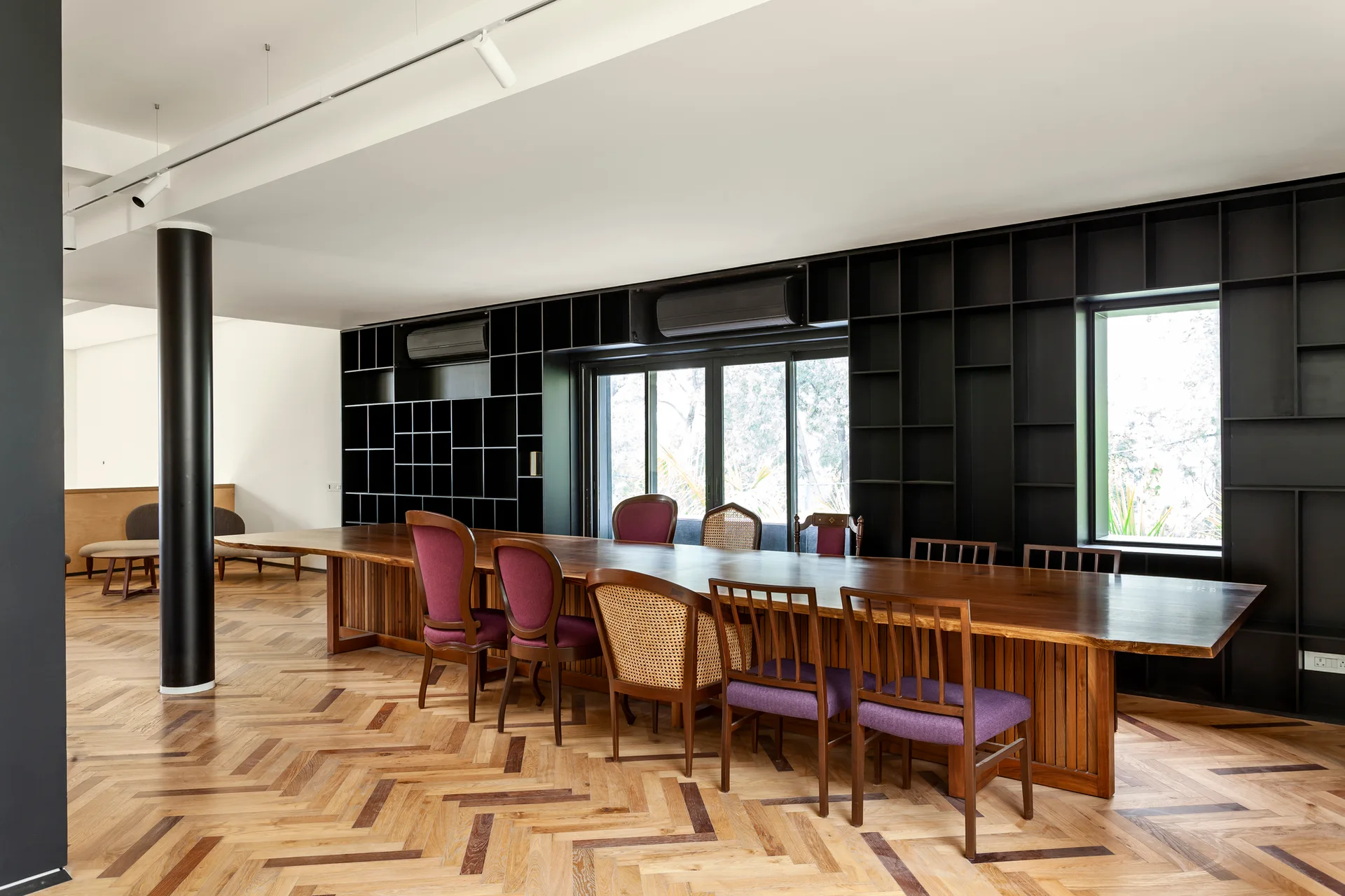 A large wooden slab dining table surrounded by mismatched chairs is set against a black grid shelving wall and herringbone wood floors.
