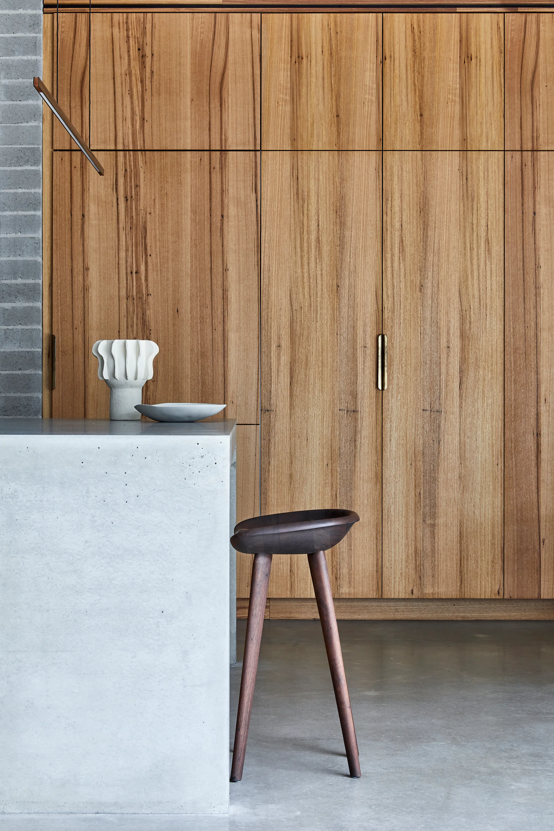 A minimalist kitchen features a grey concrete island with a dark wood stool in front of a full-height wooden storage wall.