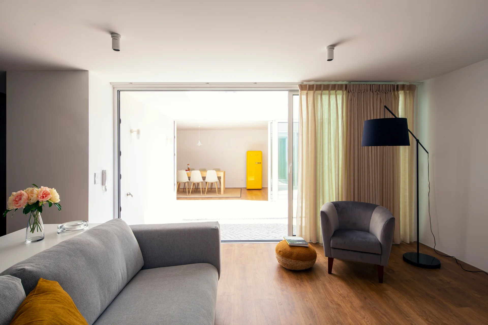 A grey sofa and armchair sit in a bright living room near sliding glass doors that look out toward a distant dining table and a yellow refrigerator.