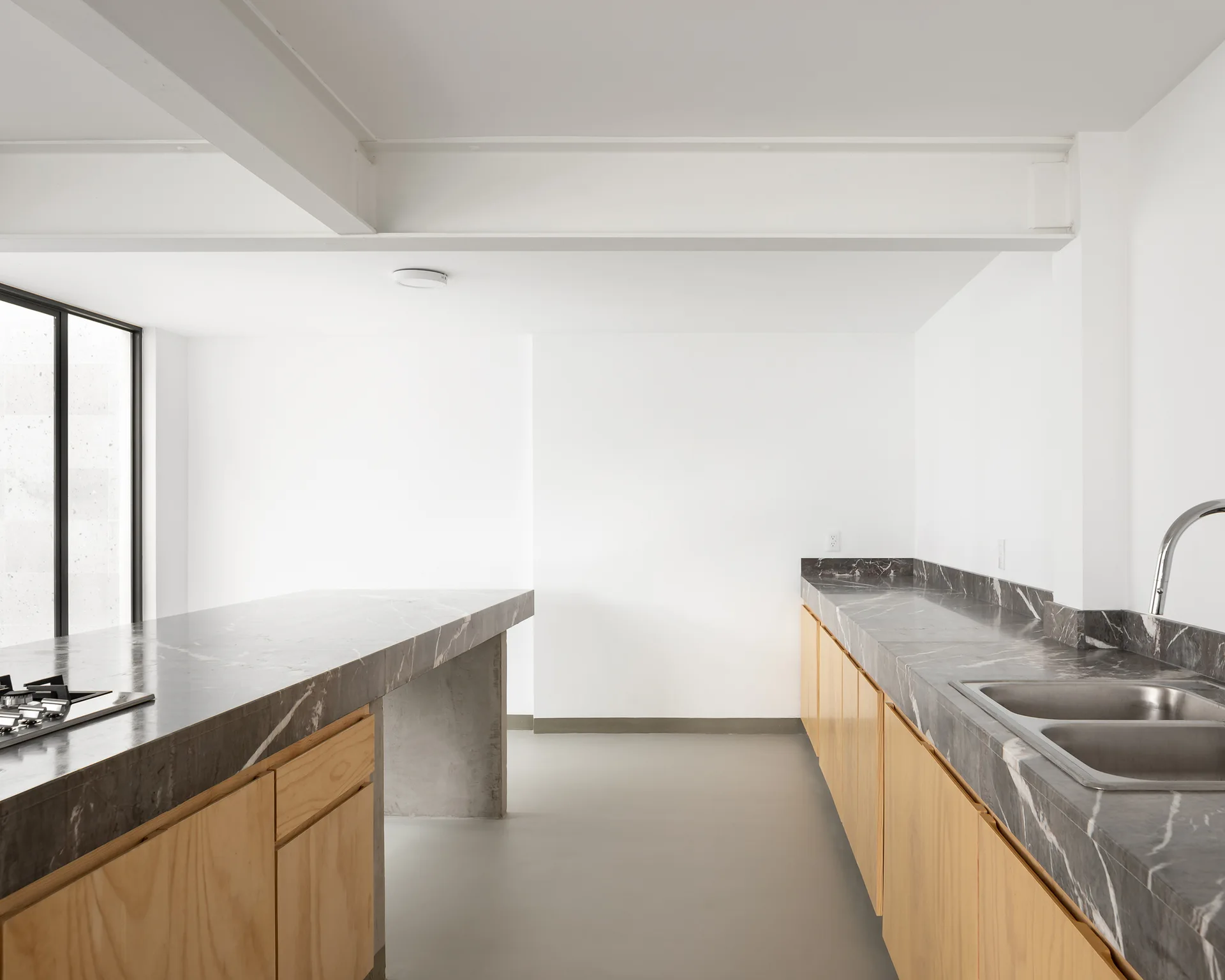 A minimalist kitchen features natural wood base cabinets, dark veined stone countertops, a double sink, and an exposed white ceiling beam.