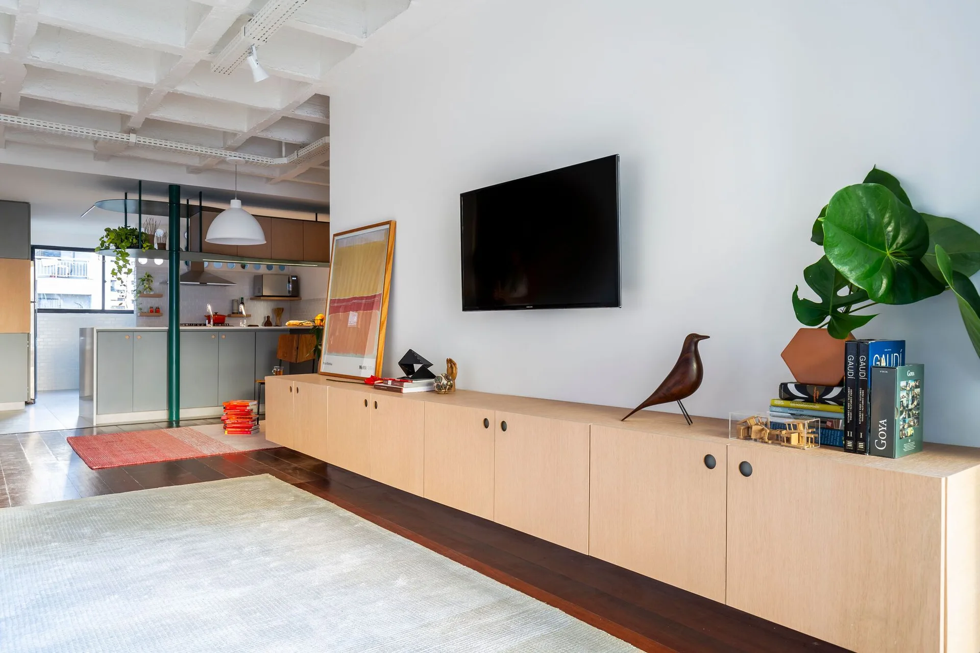 A contemporary open living room features a long light-wood media cabinet, a wall-mounted TV, and an exposed concrete waffle ceiling.
